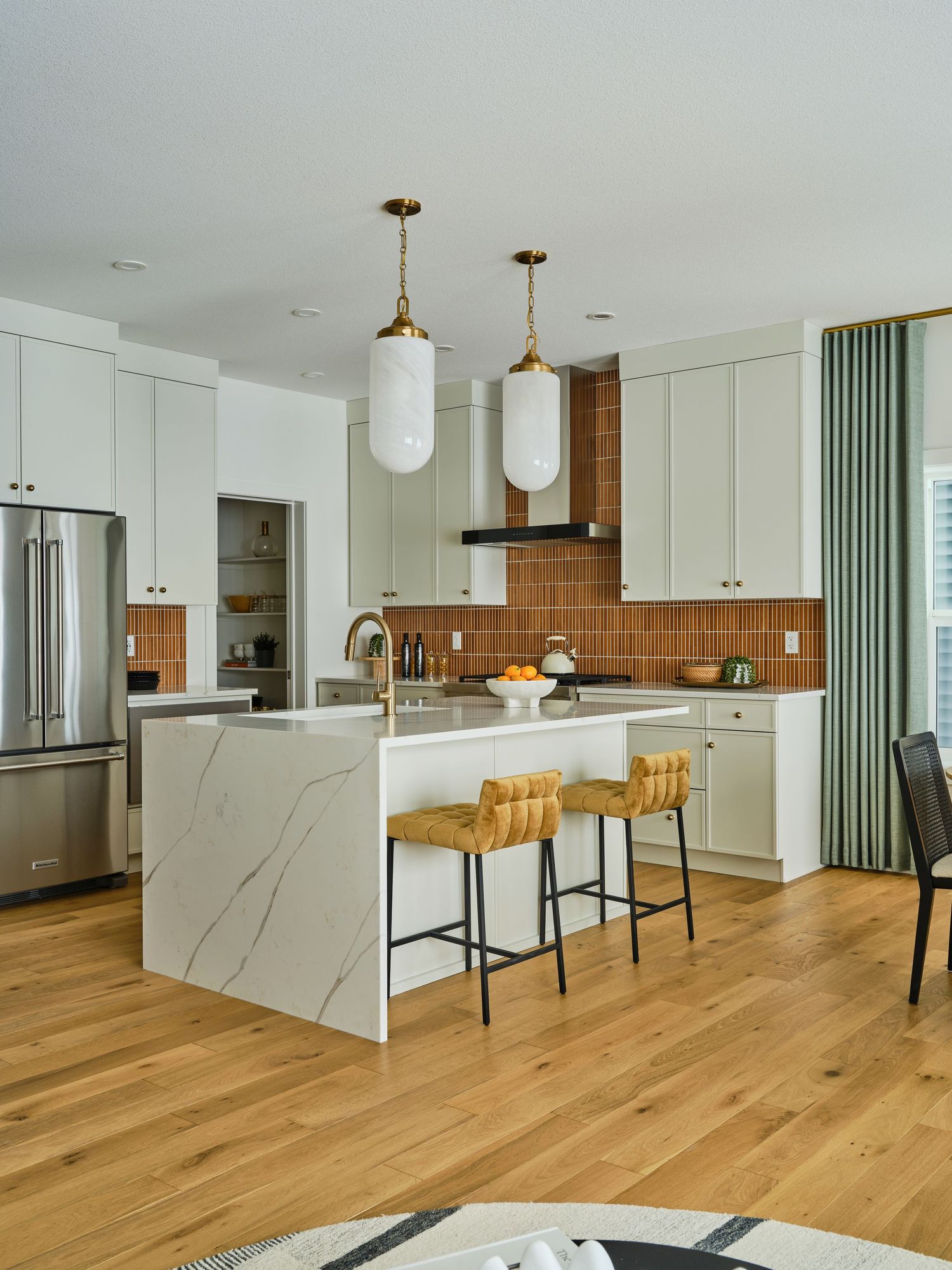 Modern white kitchen with marble island, terracotta tile backsplash, brass pendants, and woven bar stools.