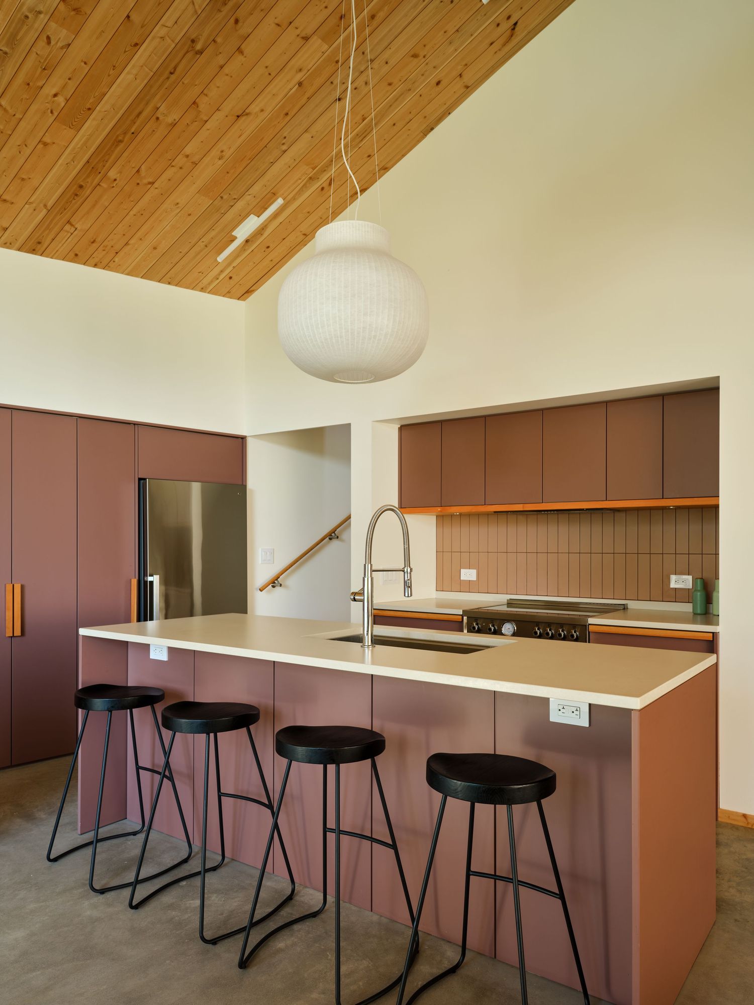 Modern kitchen with mauve cabinetry, white island, black bar stools, globe pendant light, and vaulted wood ceiling.