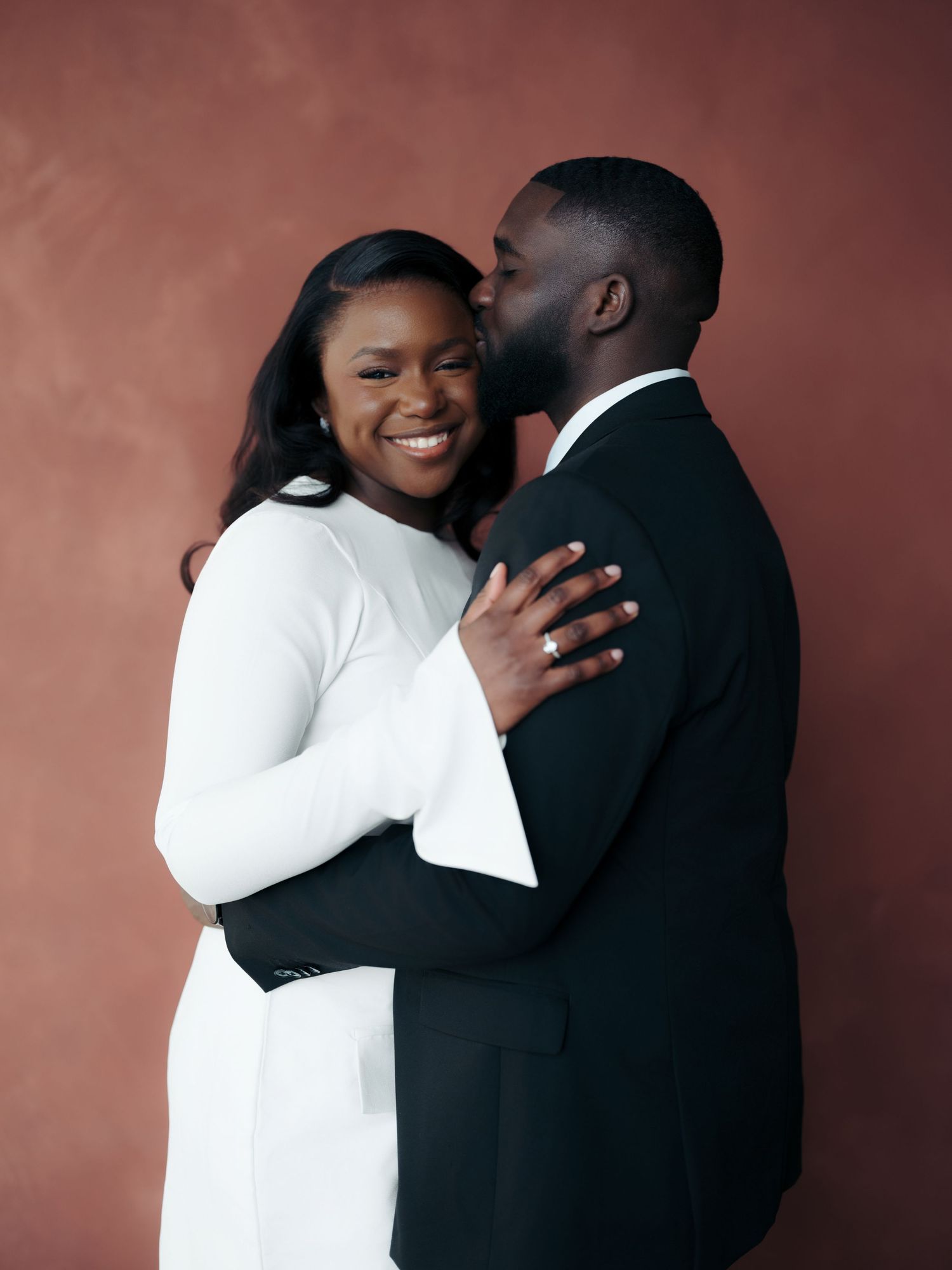Happy couple embracing and sharing a kiss in white dress and black suit against a warm terracotta wall.