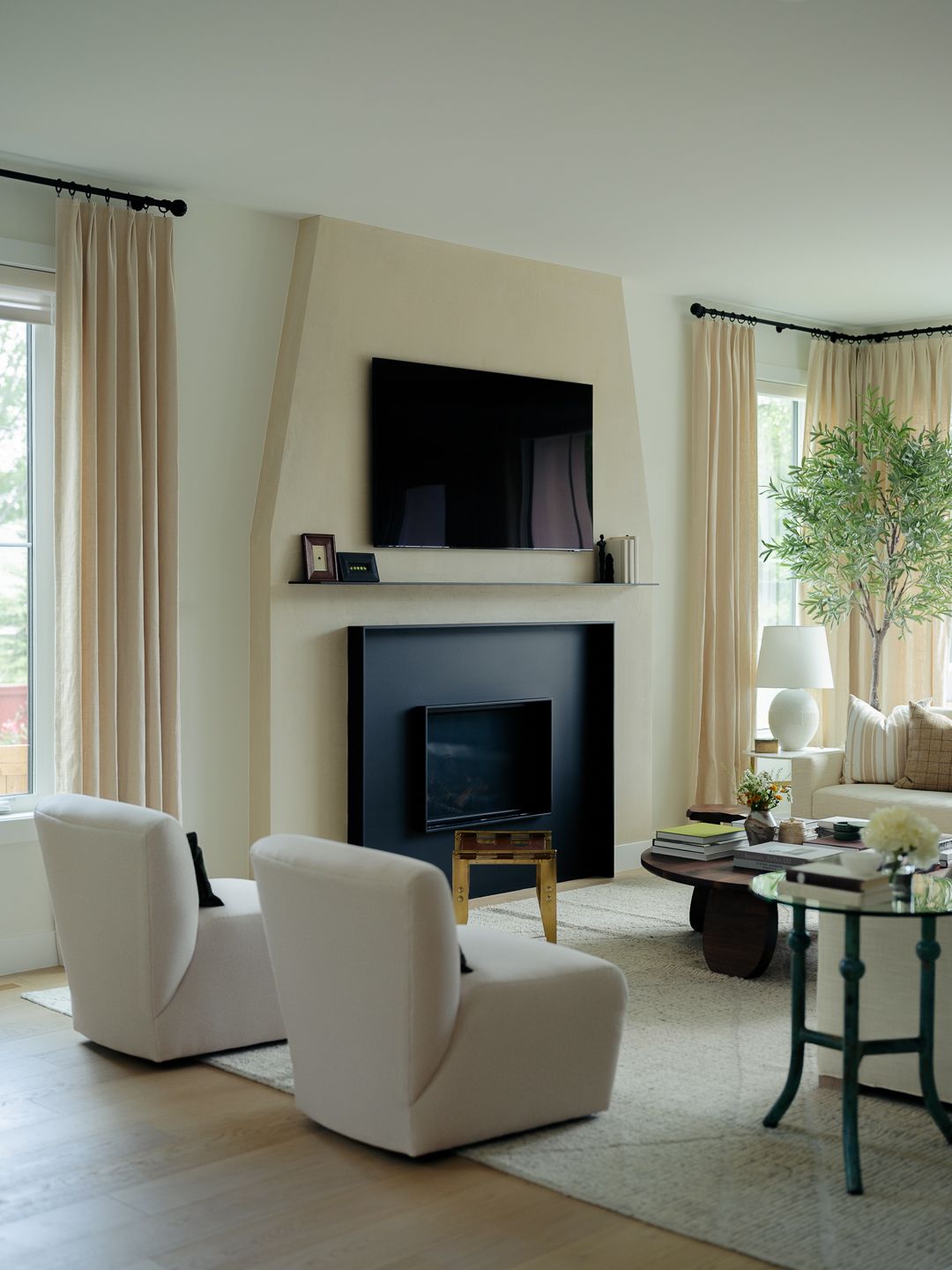 Elegant living room with white fireplace, mounted TV, cream armchairs, draped curtains, and indoor olive tree.