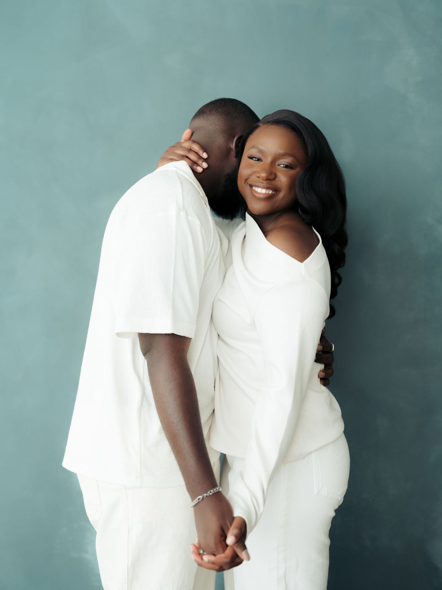 Romantic couple in matching white outfits embracing and smiling against a soft teal studio backdrop.