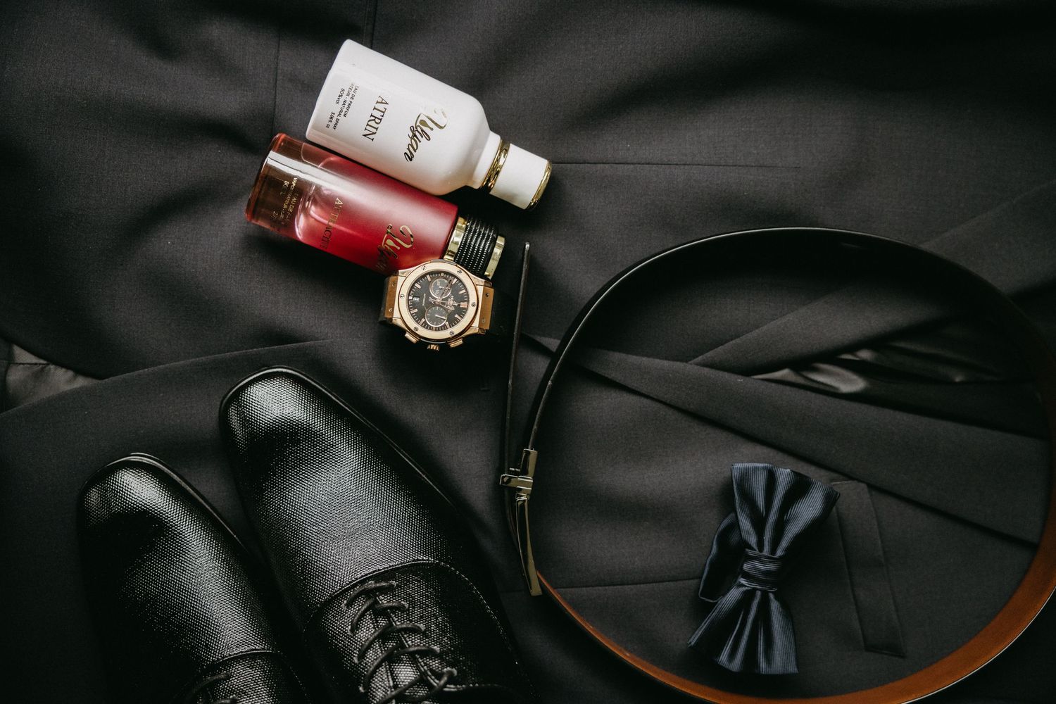 Groom wedding day details flatlay with black dress shoes, belt, watch, bow tie, and cologne.