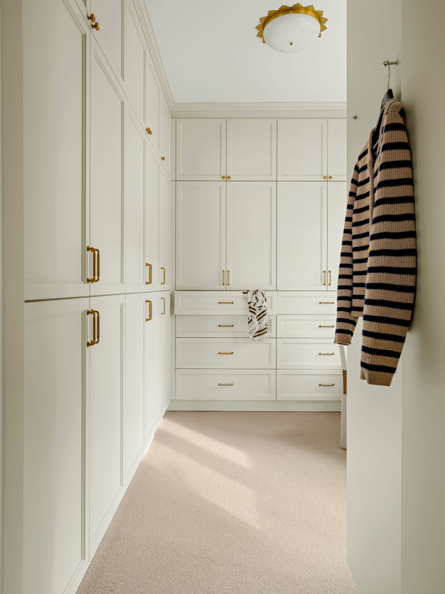Elegant walk-in closet with white shaker cabinets, brass hardware, built-in drawers, and a striped sweater hung on a wall hook.