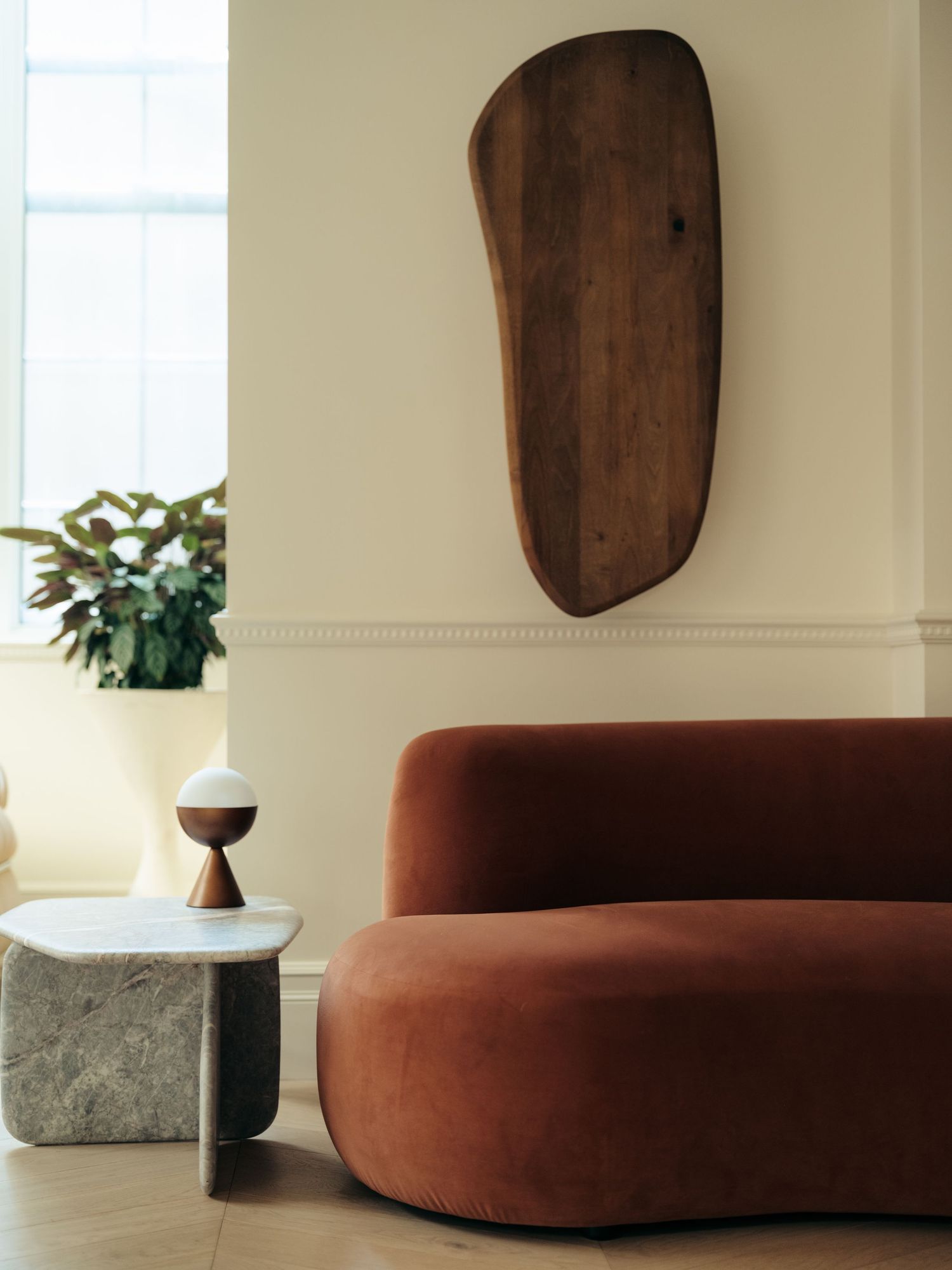 Minimalist living space featuring curved rust velvet sofa, organic wood wall art, and marble side table with globe lamp.