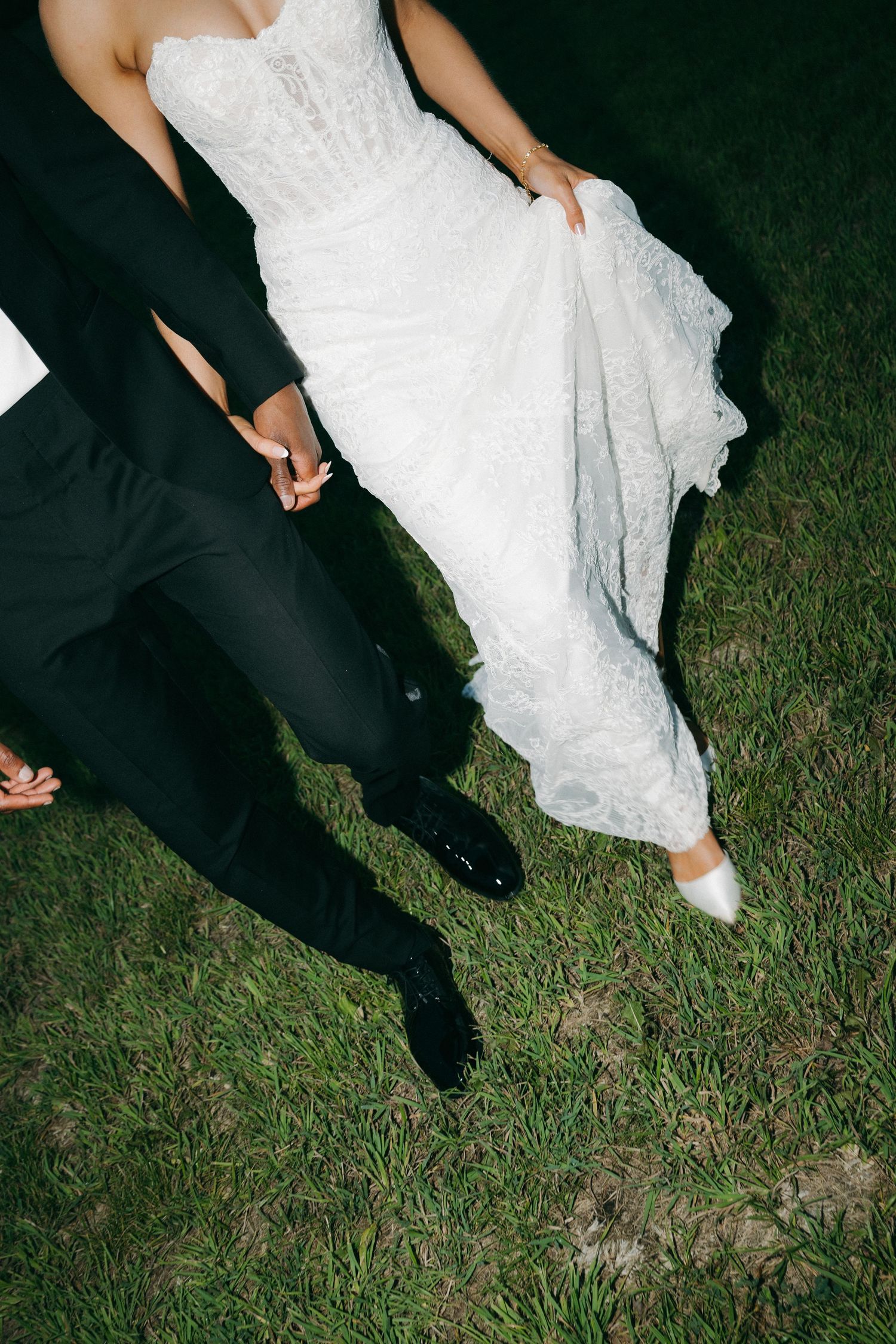 Close-up of bride in lace gown and groom in tuxedo holding hands on grass.