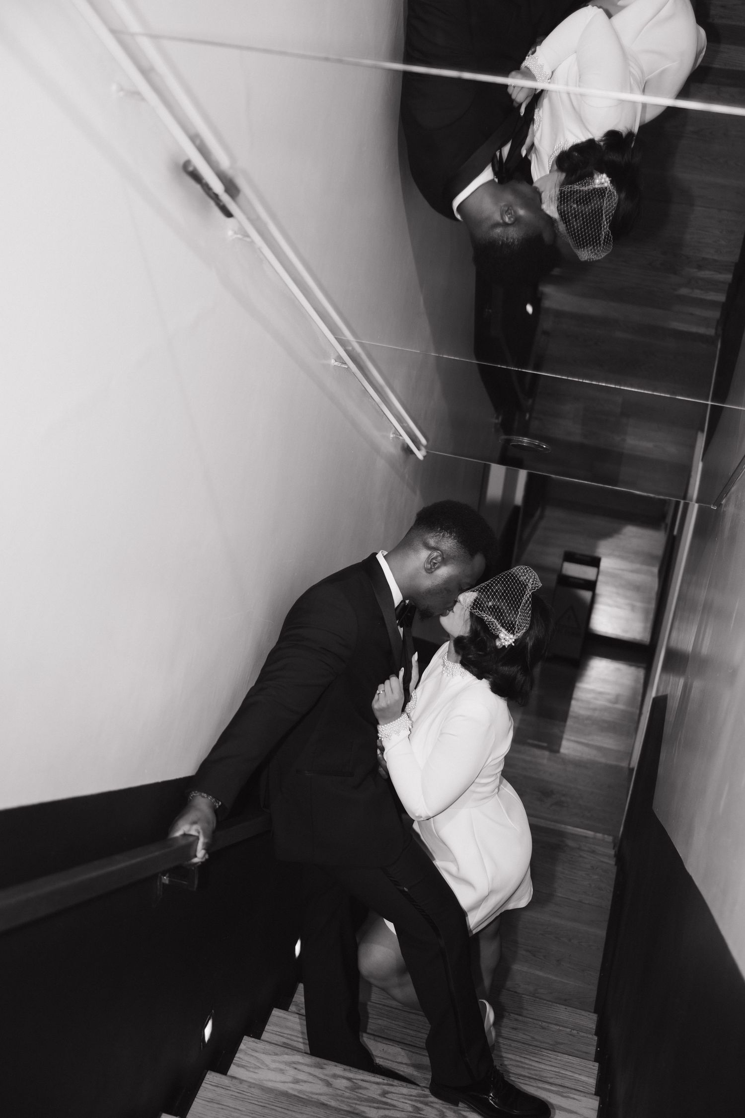 Black and white overhead photo of bride and groom sharing a kiss on modern staircase with glass railing.