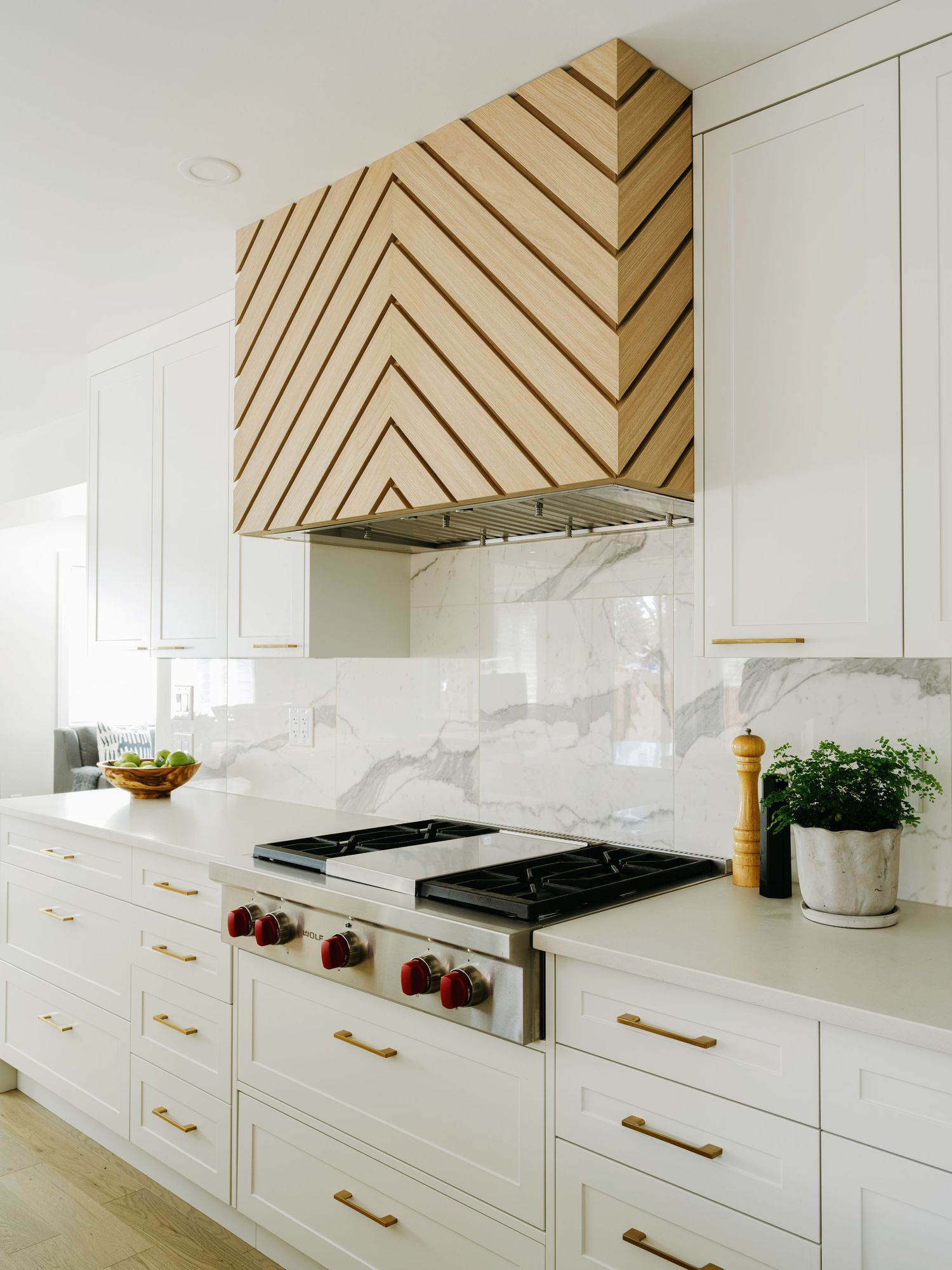 White kitchen with chevron wood range hood, marble backsplash, stainless steel range, and brass hardware.