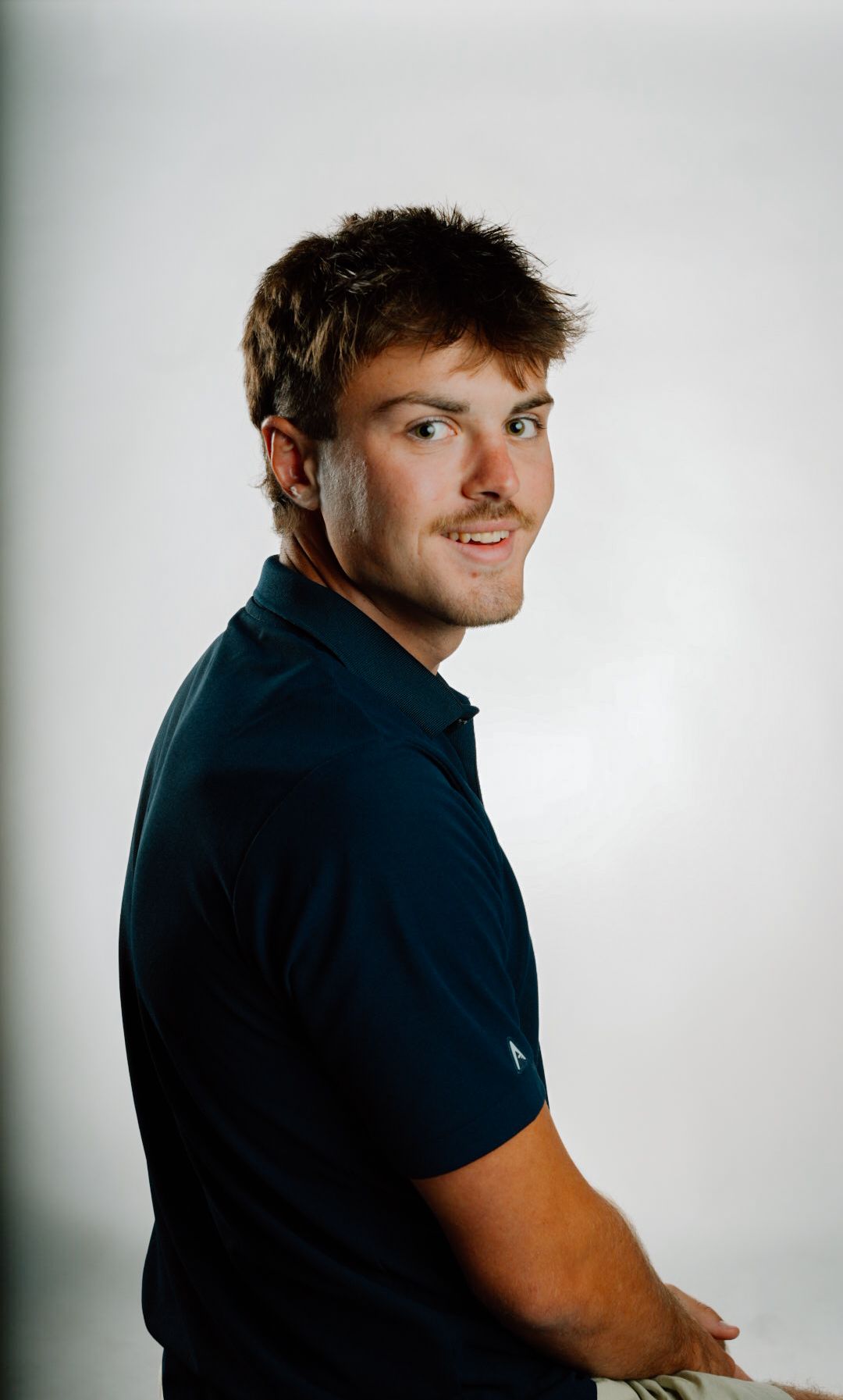 Young man with brown hair smiling over shoulder wearing navy blue polo shirt against white background.