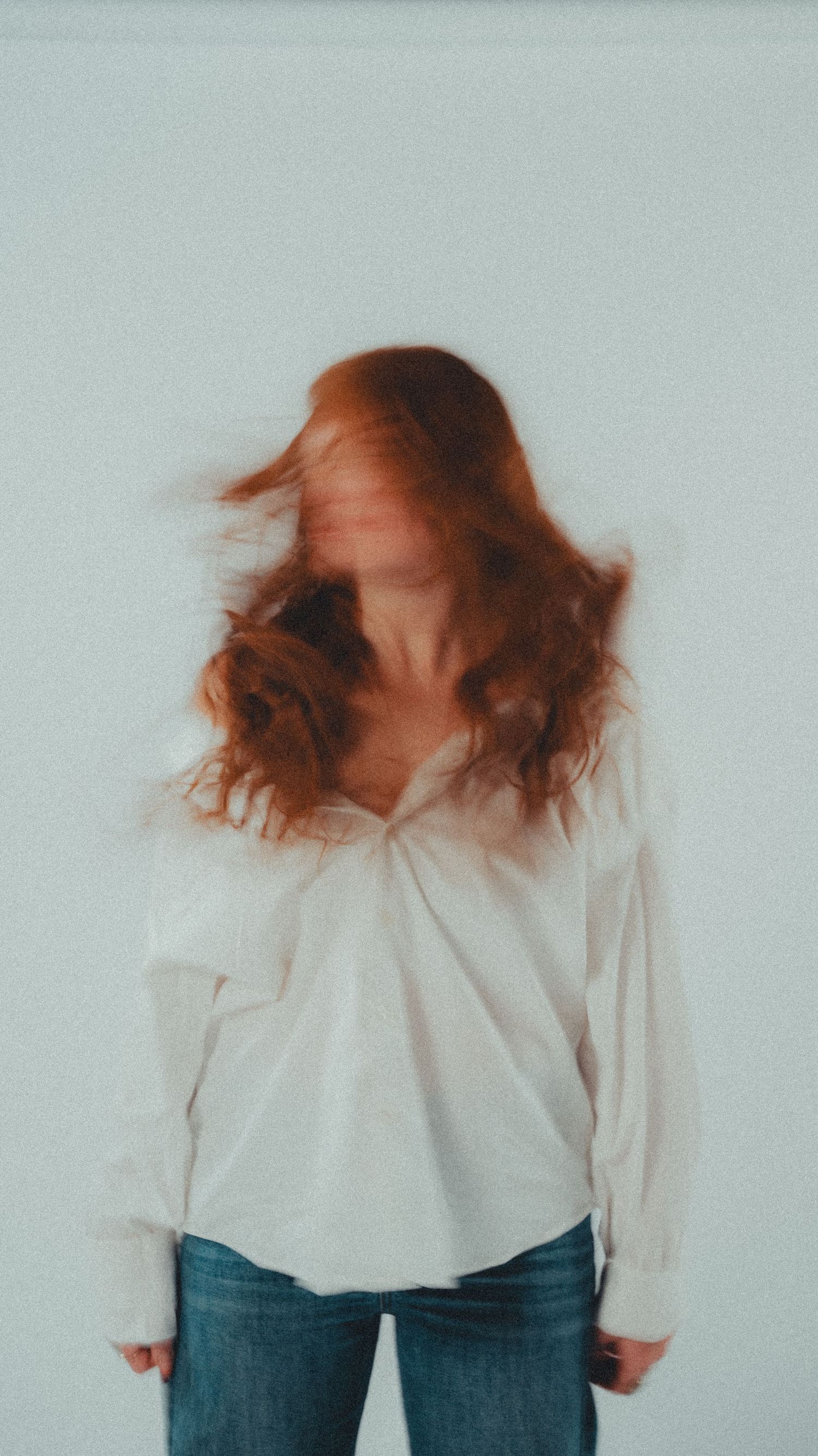 Redhead woman in white shirt and jeans with motion blur hair against light background.