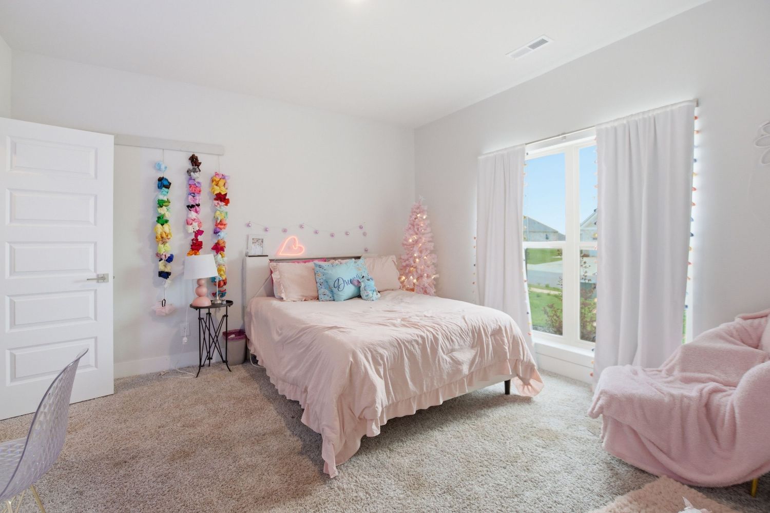 Pink-themed girl's bedroom with soft carpet, sheer curtains, floral wall decor, and a cozy bed with pastel bedding.