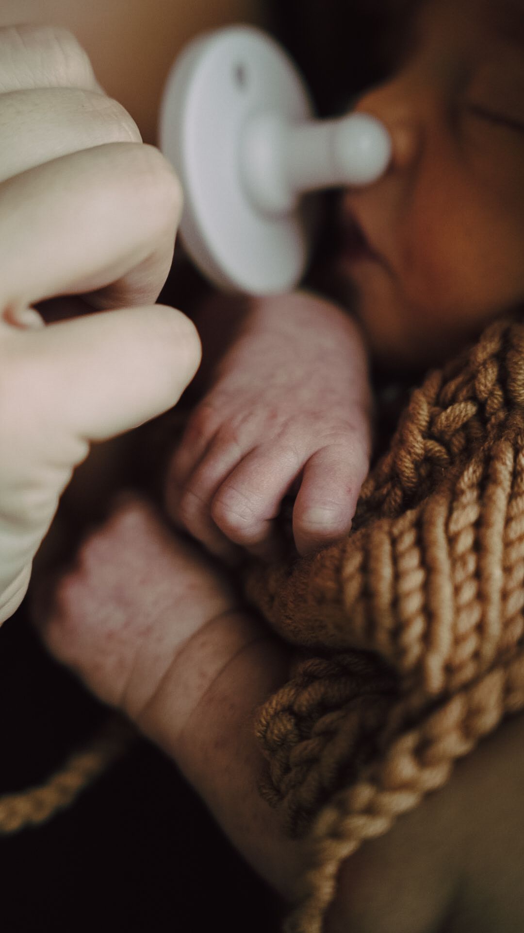 Newborn baby with pacifier gripped by gloved hand, wrapped in knit blanket, tiny fingers visible.