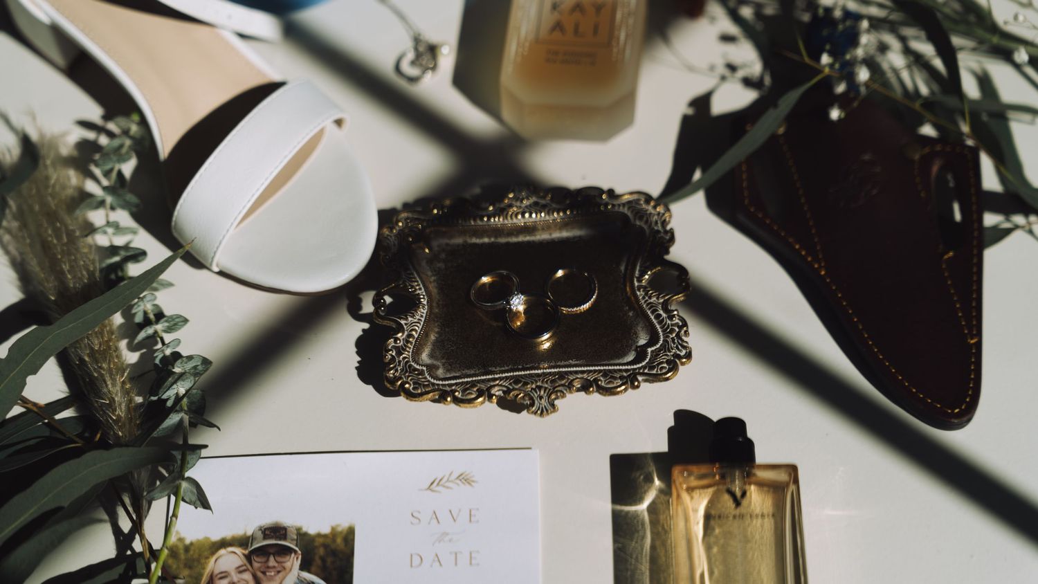 Elegant wedding flat lay featuring bridal heels, gold rings on ornate tray, save the date card, and floral accents.