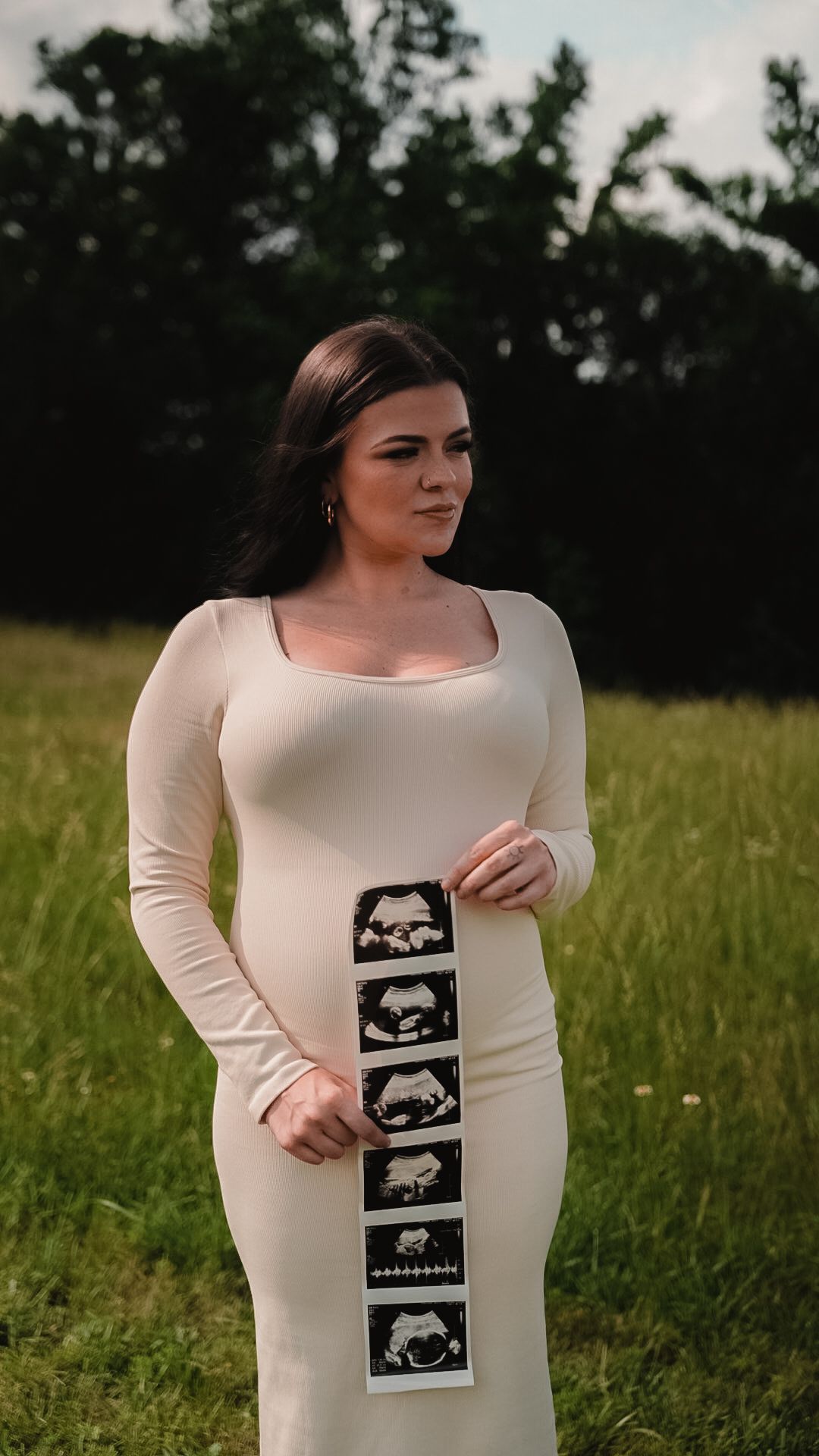 Pregnant woman in white dress holding ultrasound strip photos while standing in green field.
