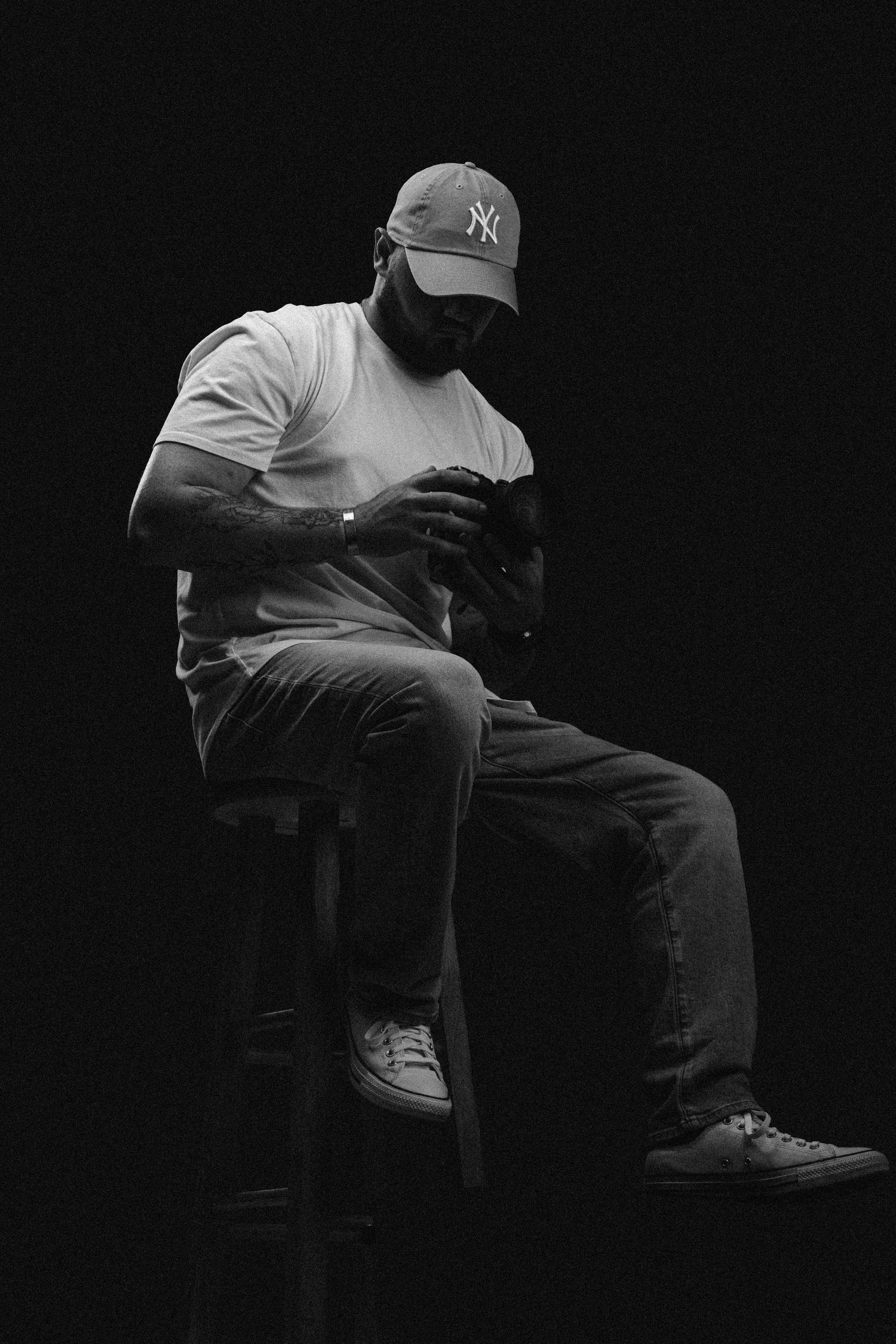Man in NY Yankees cap sits on stool looking down, dramatic black and white portrait photography.