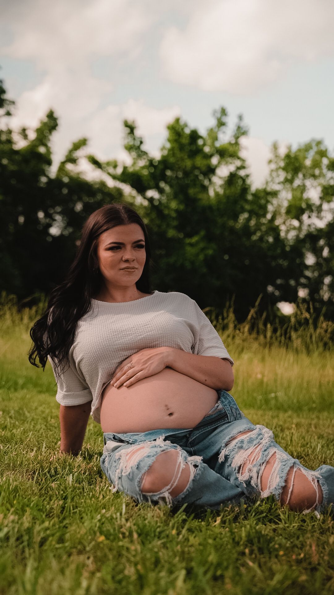 Pregnant woman sitting on grass in ripped jeans and white crop top, cradling baby bump outdoors.