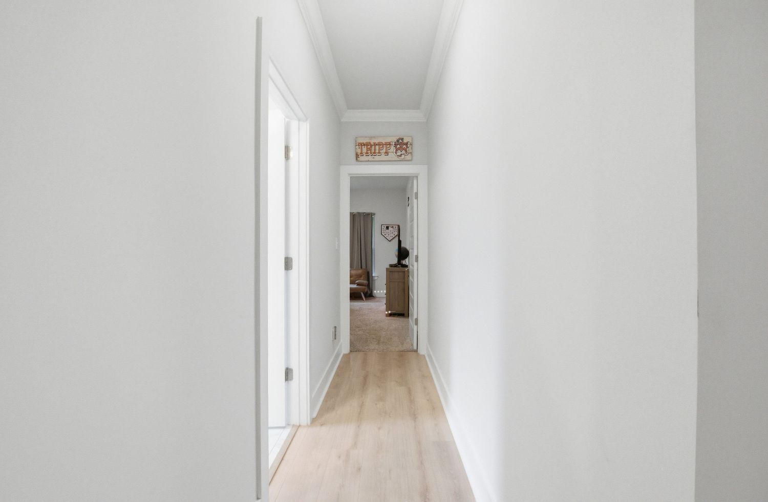 Bright narrow hallway with white walls, white doors, and light hardwood floors leading to a warmly decorated room.