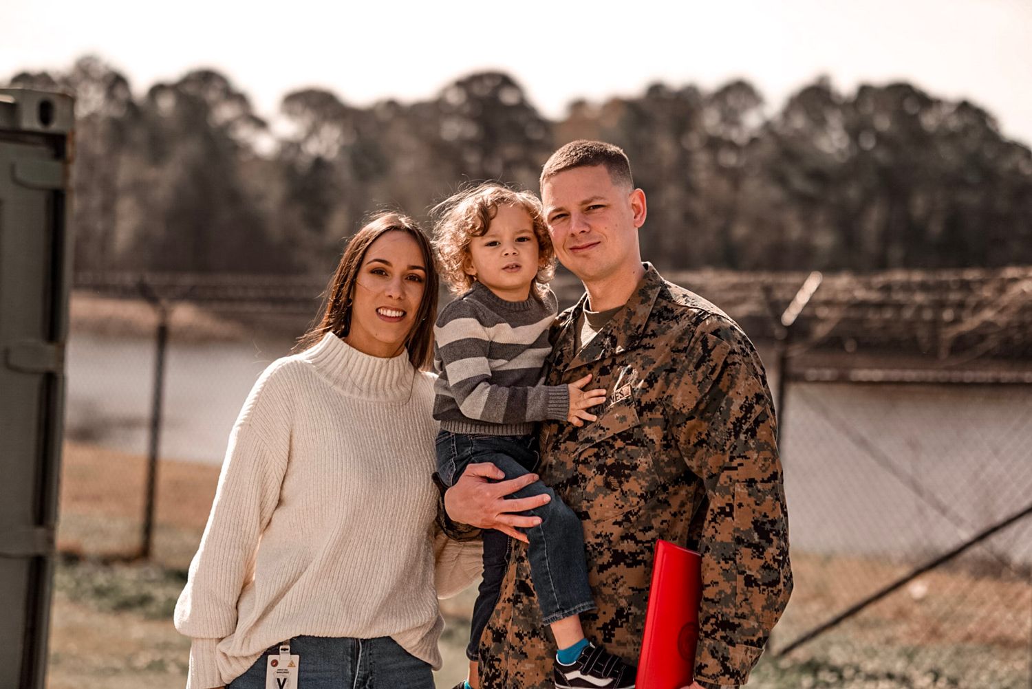 Smiling military family with young toddler posing outdoors at a base in camouflage uniform.