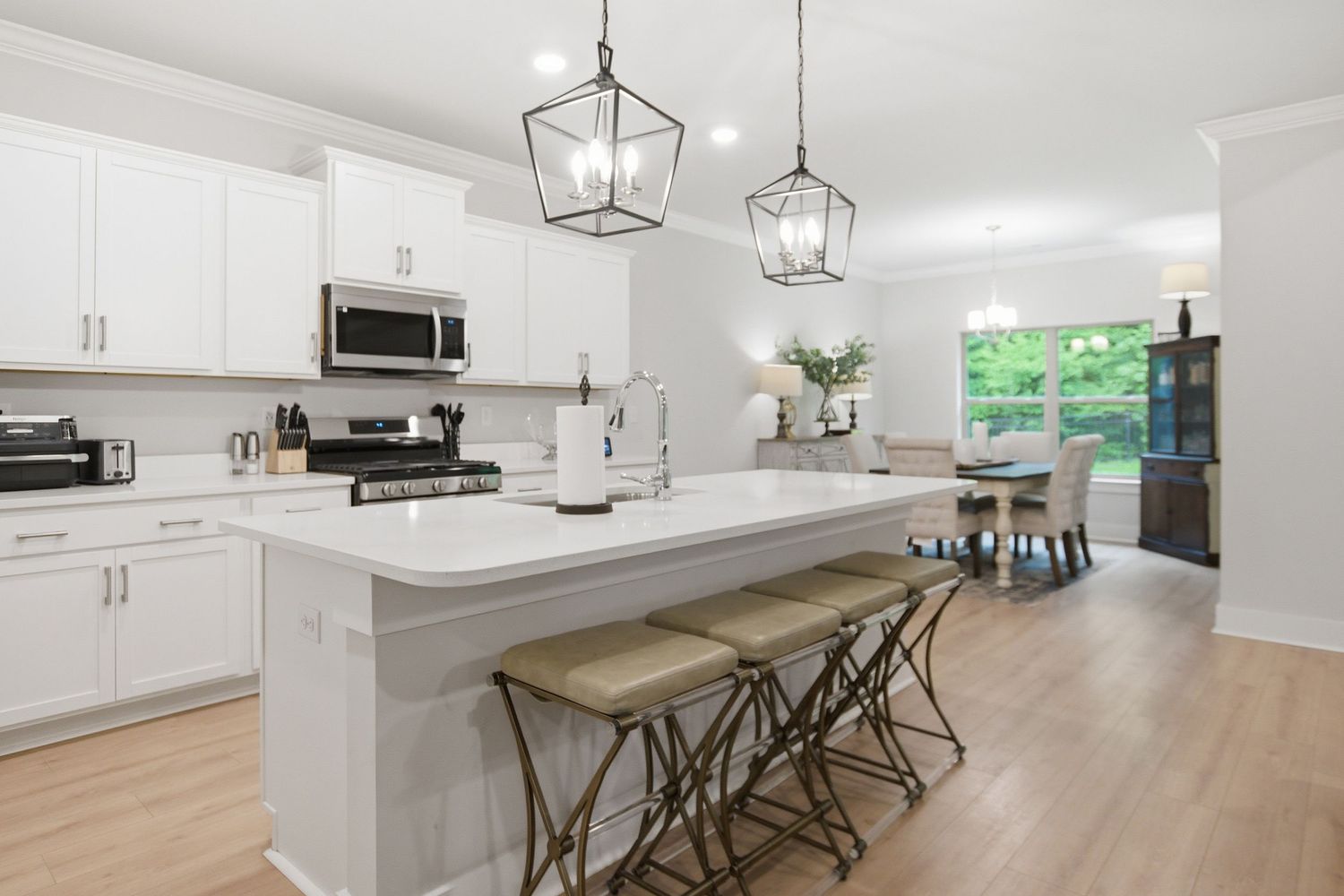 Modern white kitchen with island, bar stools, pendant lights, stainless appliances, and open dining area with natural light.