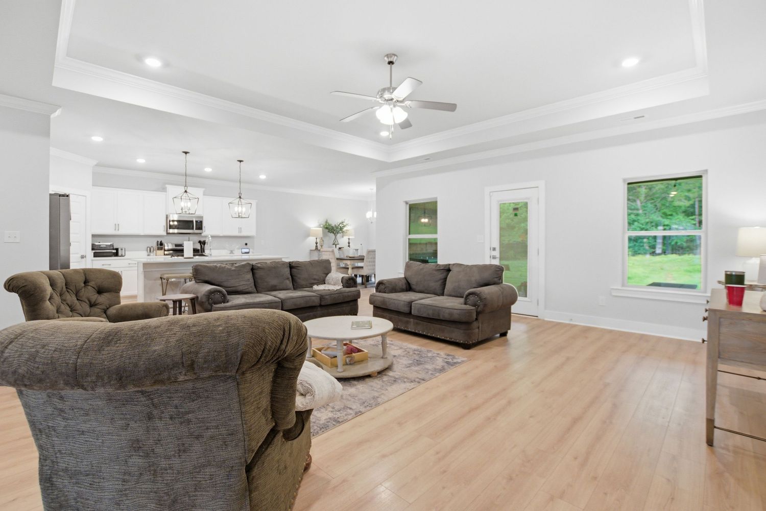 Spacious open-concept living room with hardwood floors, gray sofas, tray ceiling, ceiling fan, and modern white kitchen in background.