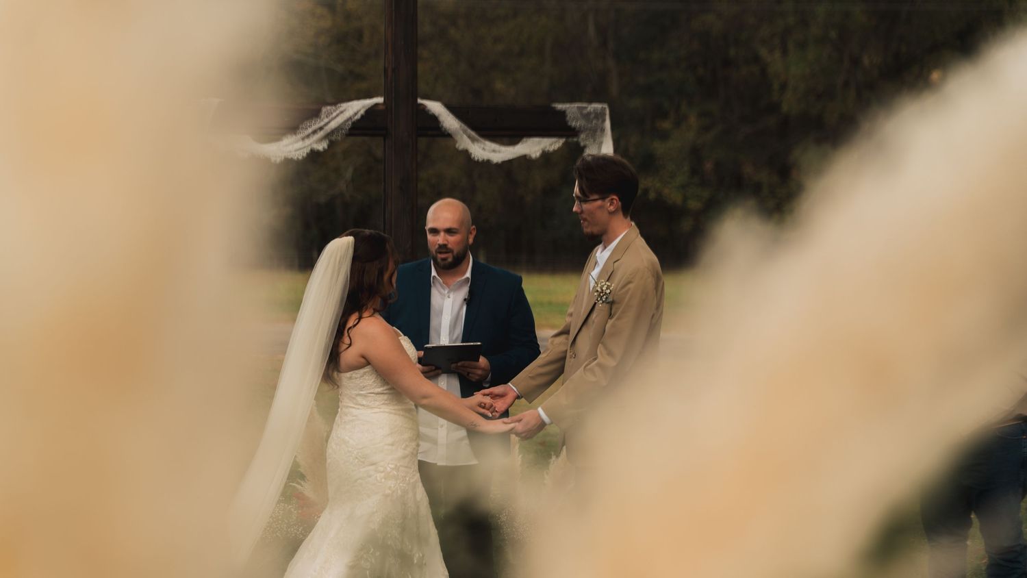 Outdoor wedding ceremony with bride and groom holding hands before officiant, framed by soft bokeh foliage.