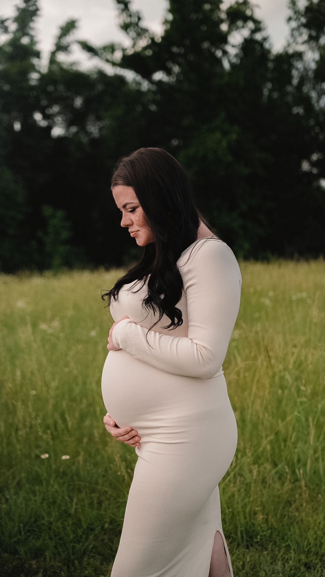 Pregnant woman in white ribbed maxi dress cradling baby bump in lush green field during maternity photo session.