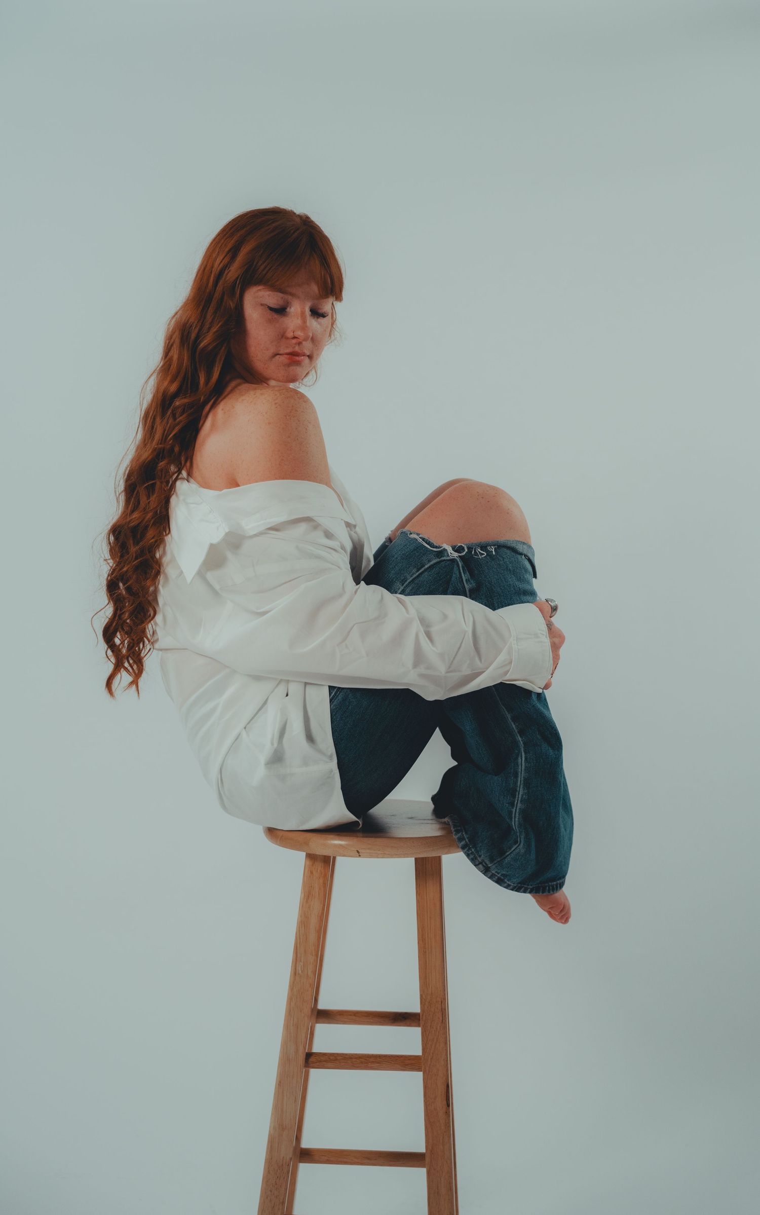 A woman with long red hair sits curled up on a wooden stool, wearing a white off-shoulder top and ripped blue jeans.
