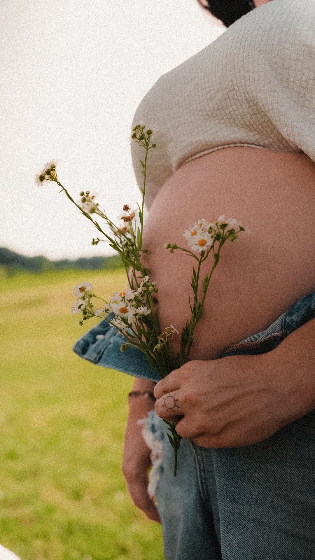 Pregnant woman holding wildflowers against her baby bump outdoors in a green field.