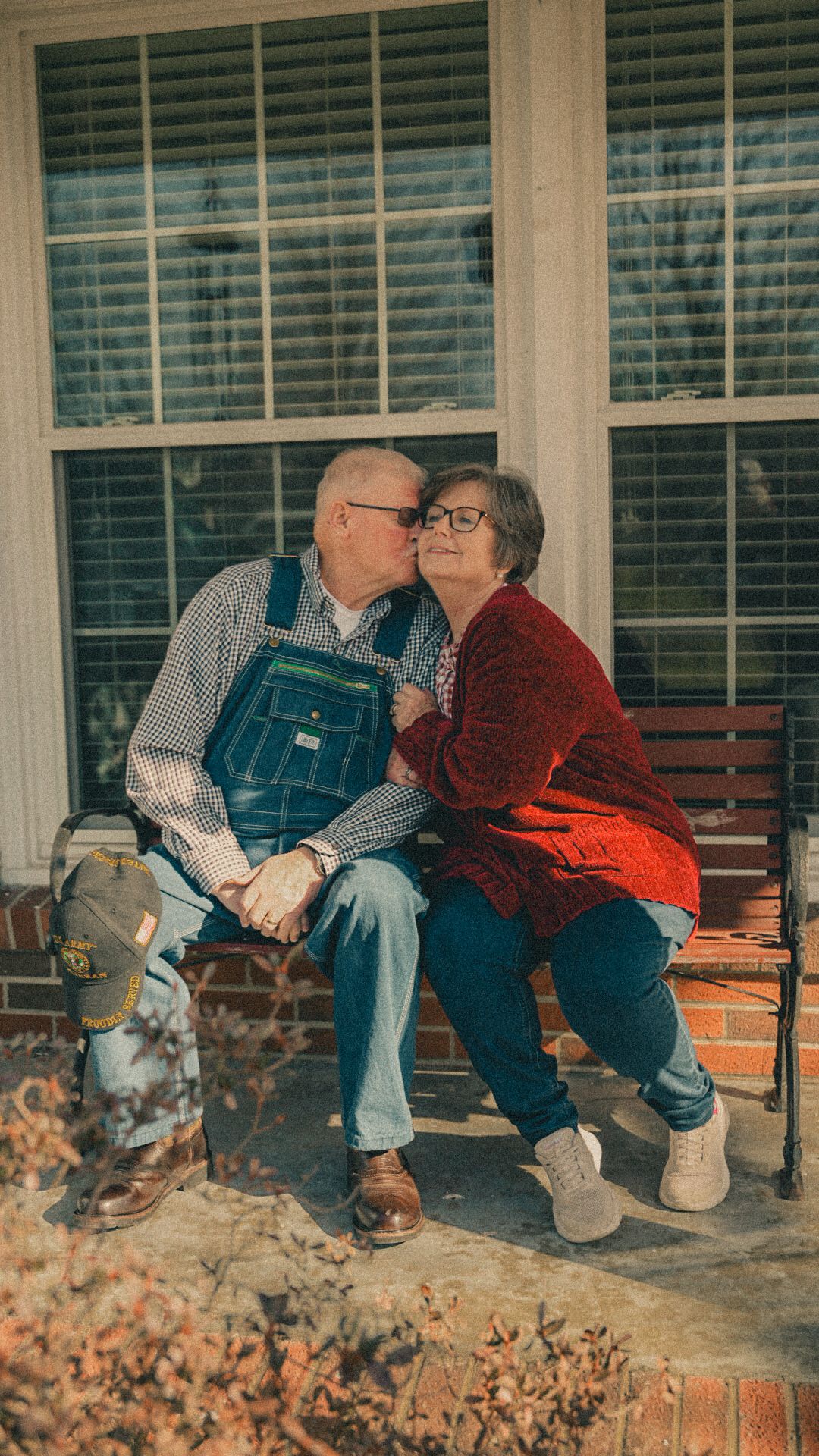 Elderly couple sharing a kiss on a porch bench, man in overalls and woman in red cardigan, holding hands.
