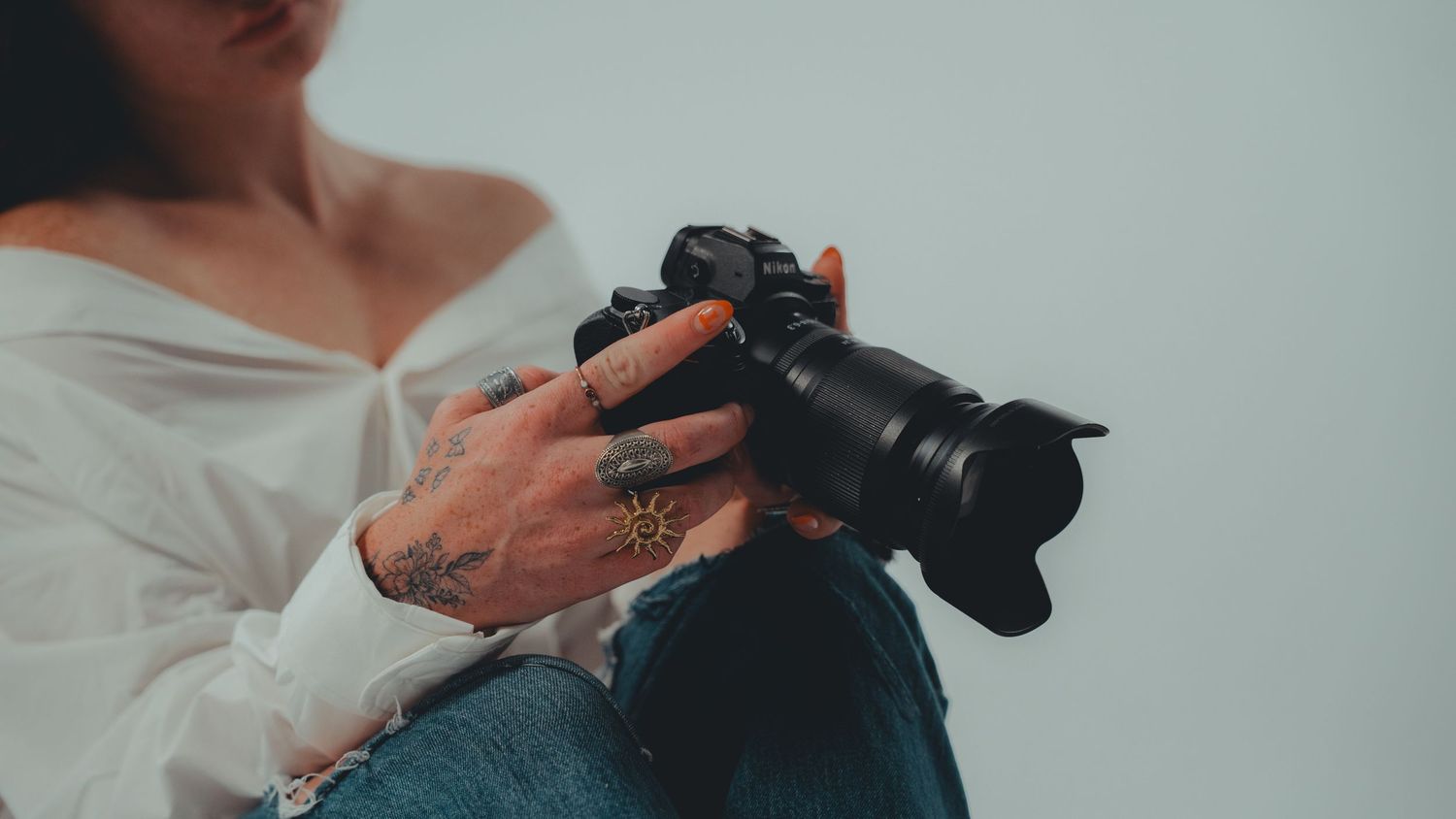 Tattooed photographer holding a professional DSLR camera, wearing white off-shoulder top and jeans.