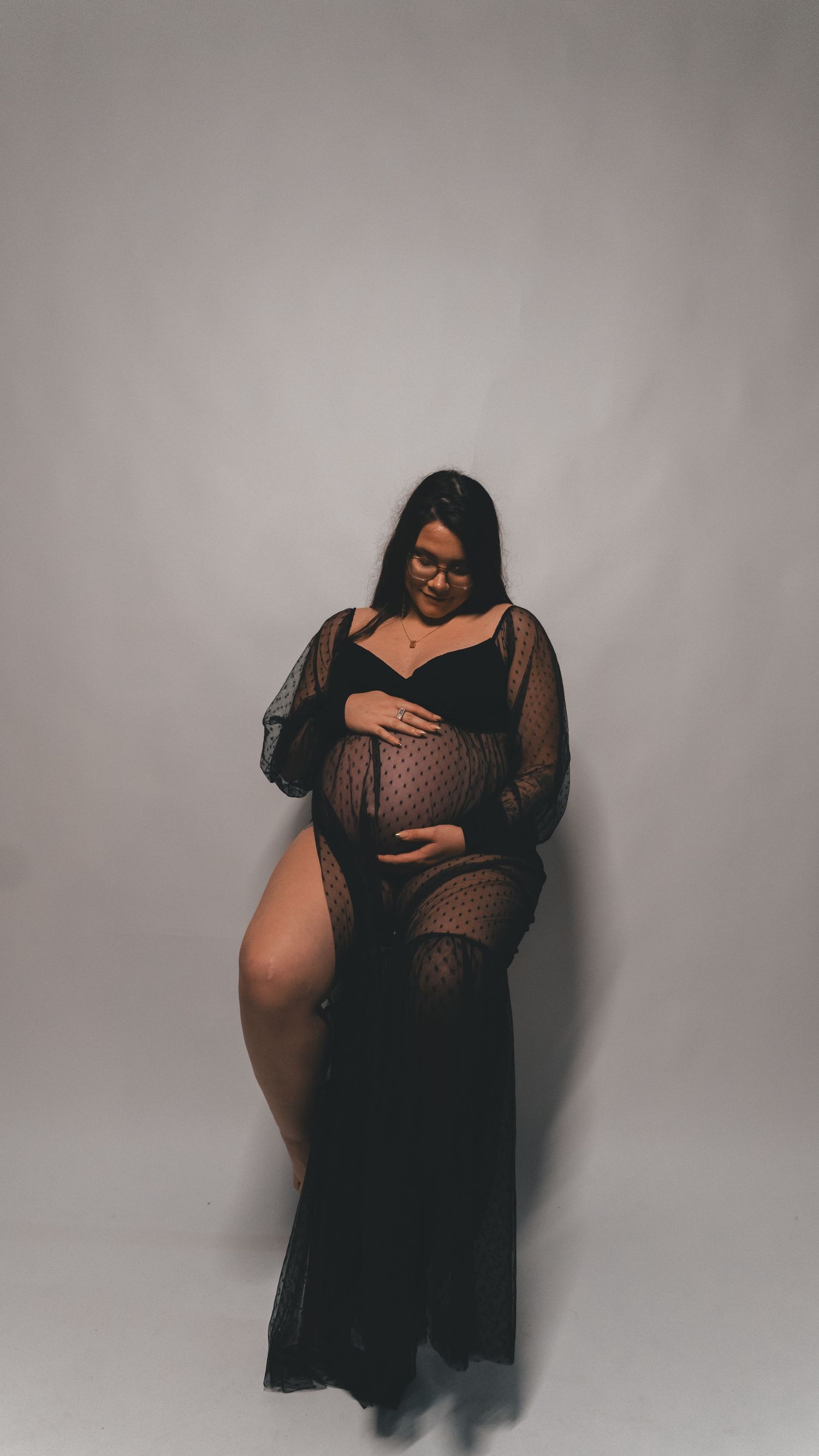 Pregnant woman in black sheer maternity gown cradling baby bump during elegant studio maternity photoshoot.