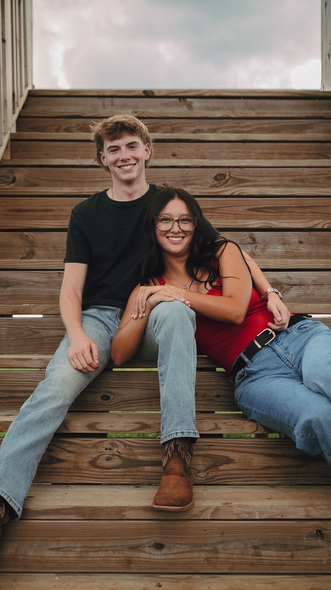 A young couple smiles while sitting together on wooden steps, wearing casual jeans and tops outdoors.