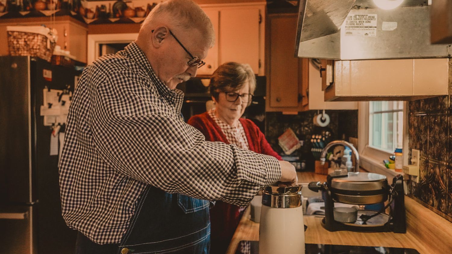 Elderly couple cooking together in a cozy kitchen, man pouring coffee while woman watches nearby.