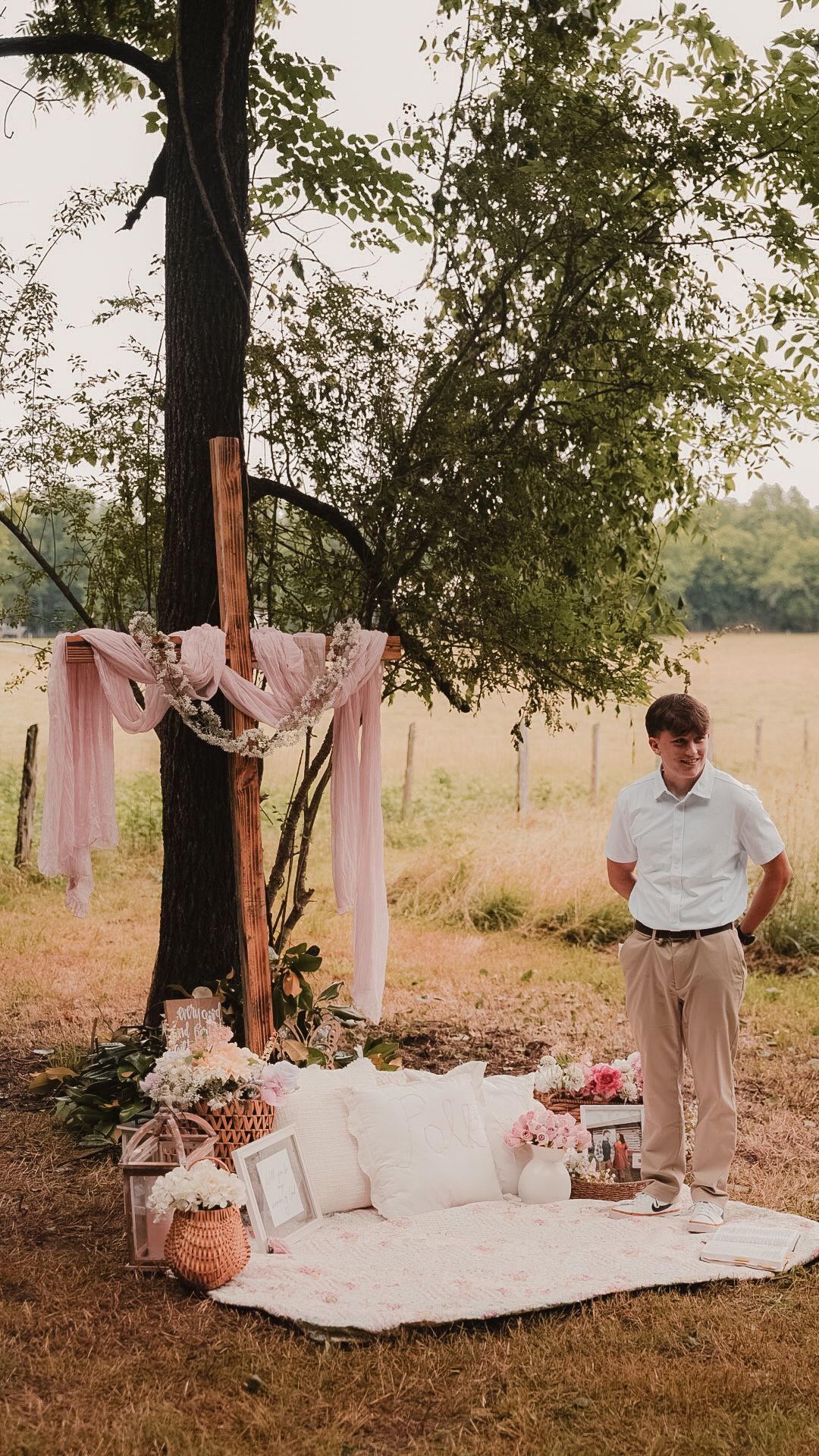 Man standing beside a romantic outdoor picnic setup with pink draping, floral decor, and a blanket under a tree in a field.
