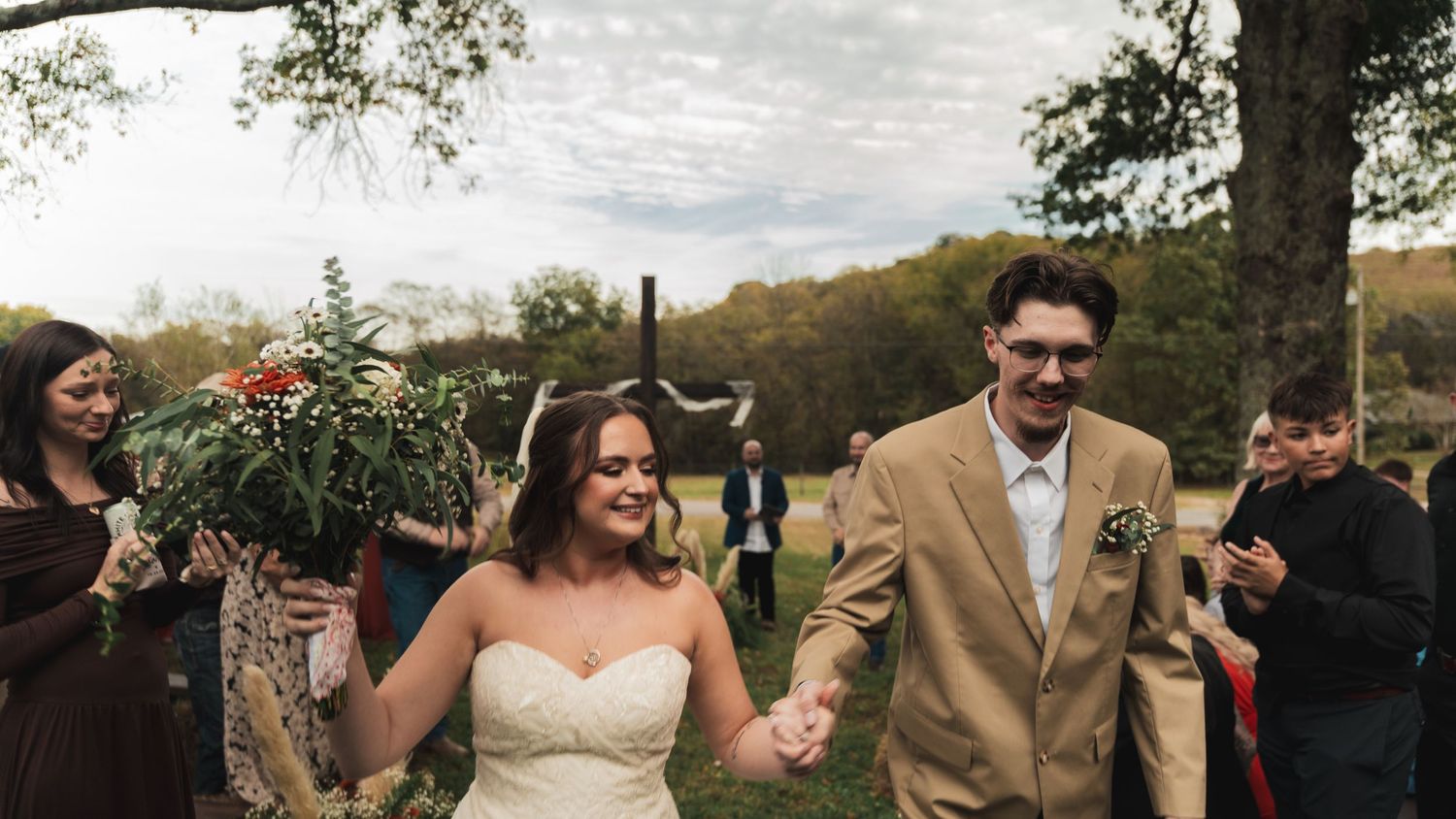 Happy newlyweds walk hand-in-hand at outdoor rustic wedding ceremony surrounded by cheering guests in autumn setting.