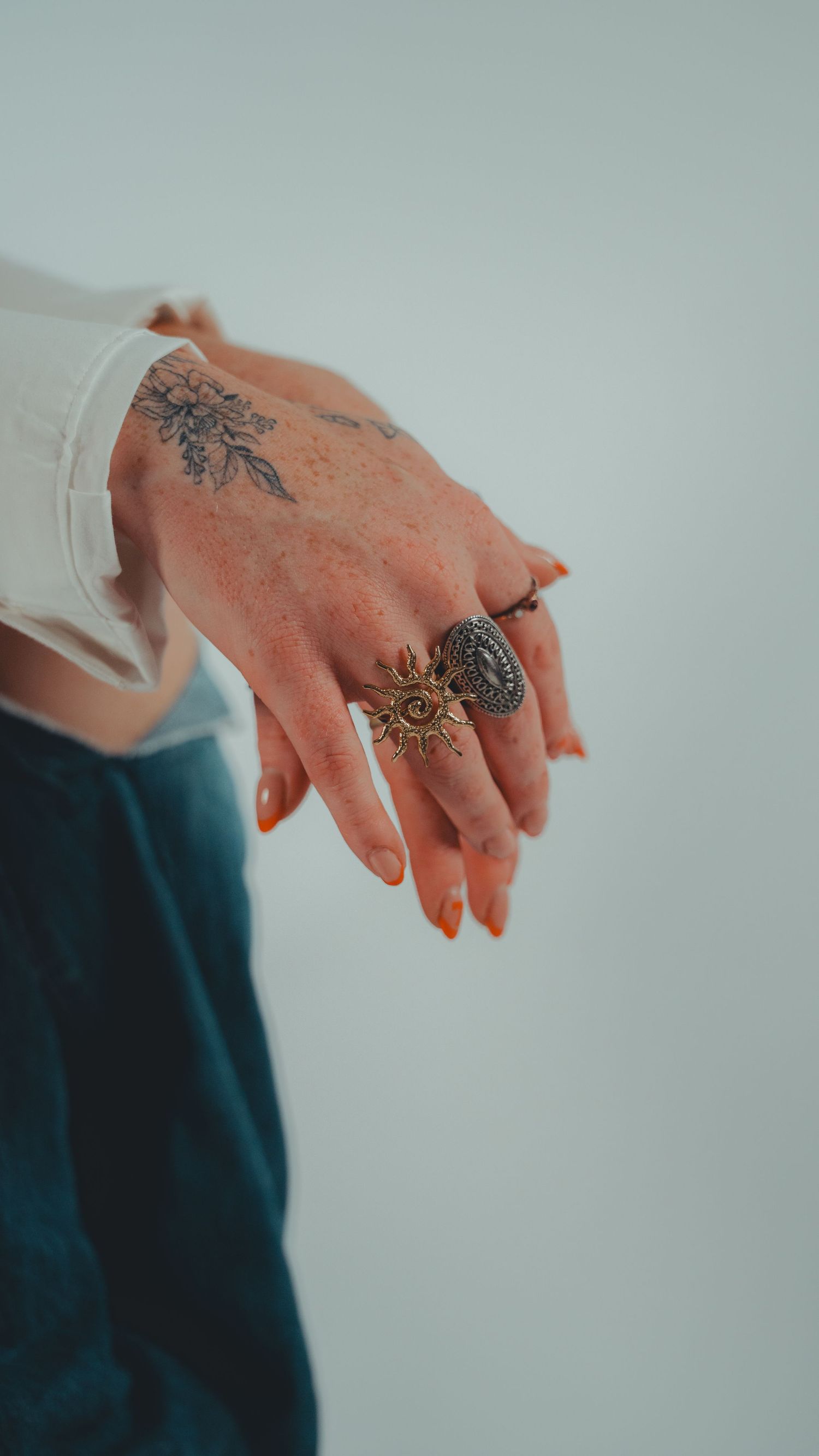 Tattooed hand with orange nails wearing gold sun and silver rings, white sleeve and blue jeans background.