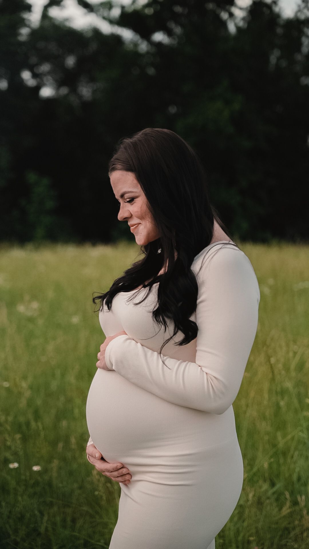 Smiling pregnant woman in white dress cradling baby bump in a green field during golden hour maternity photoshoot.