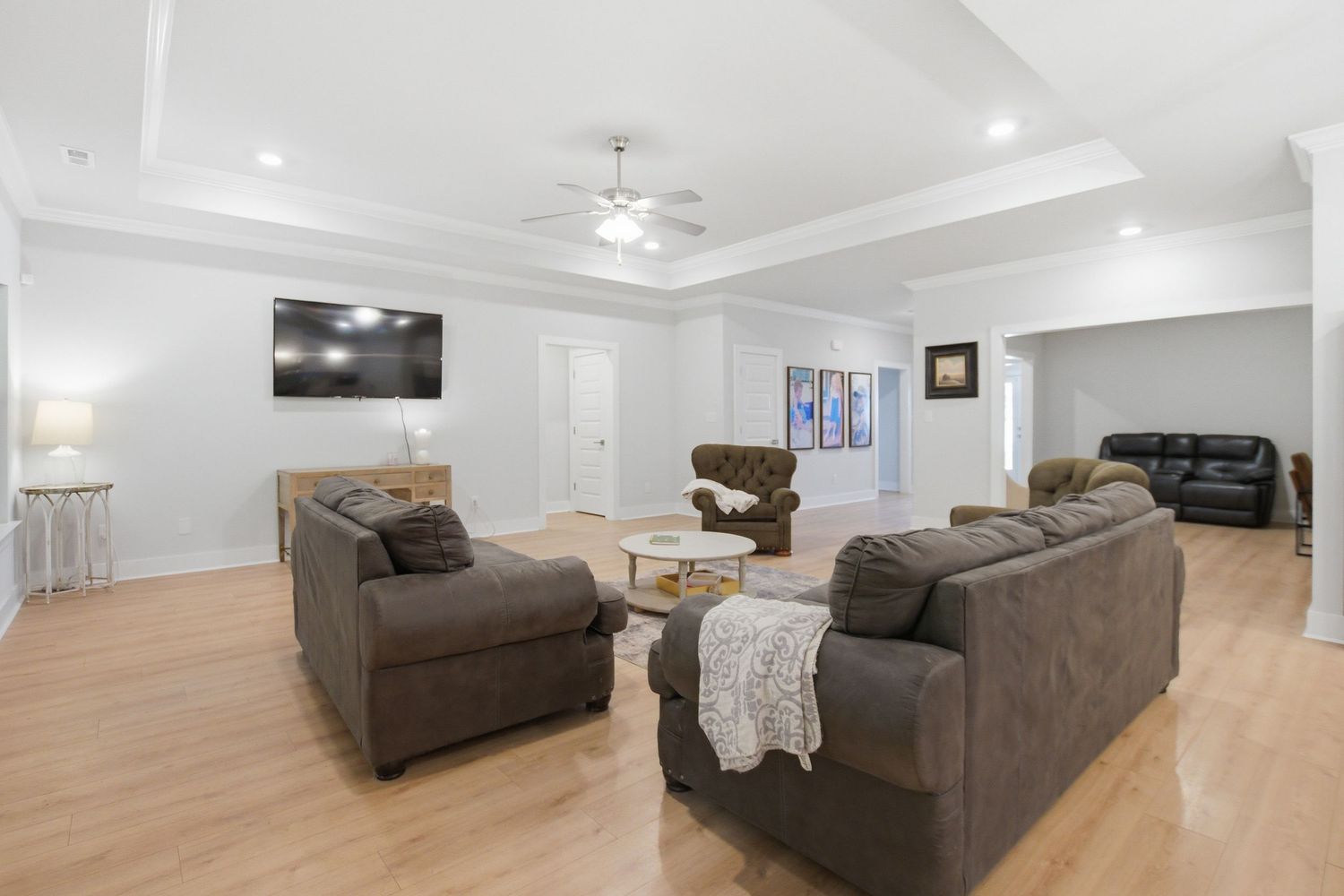 Spacious living room with brown sofas, hardwood floors, tray ceiling, ceiling fan, and wall-mounted TV in a bright home.