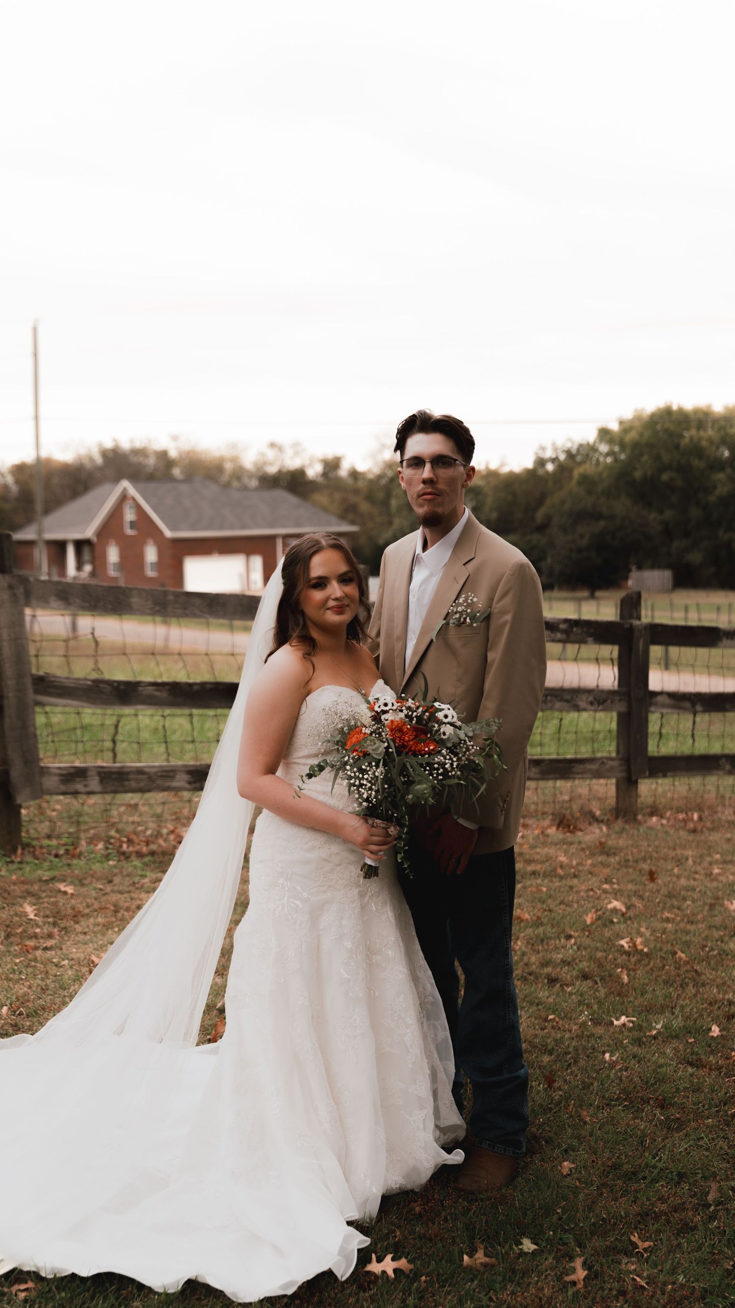 A bride in a white gown holding an autumn bouquet stands with a groom in a tan blazer at a rustic farm wedding.