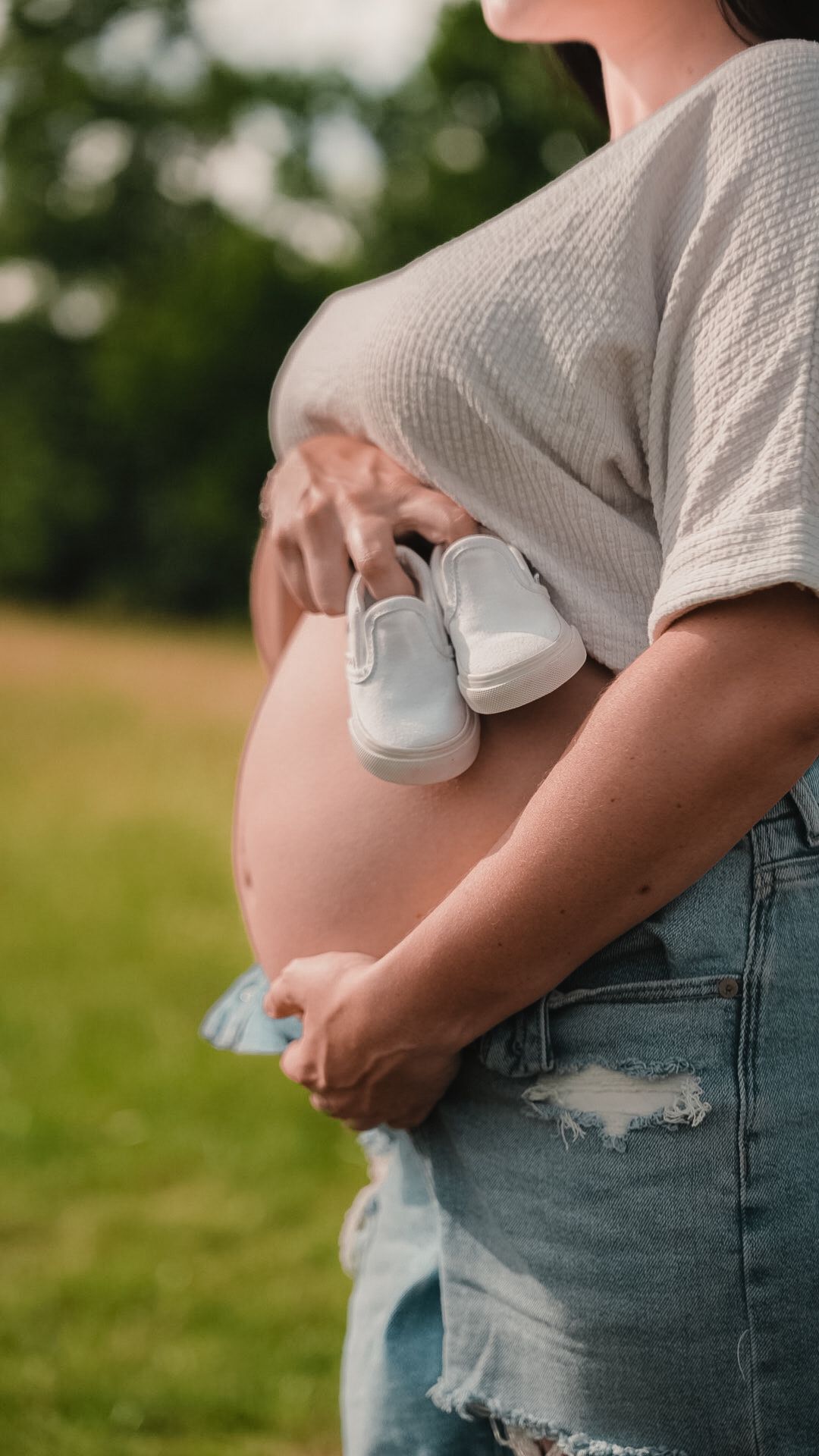 Pregnant woman in white top and ripped jeans holding tiny white baby shoes against her bare bump outdoors.