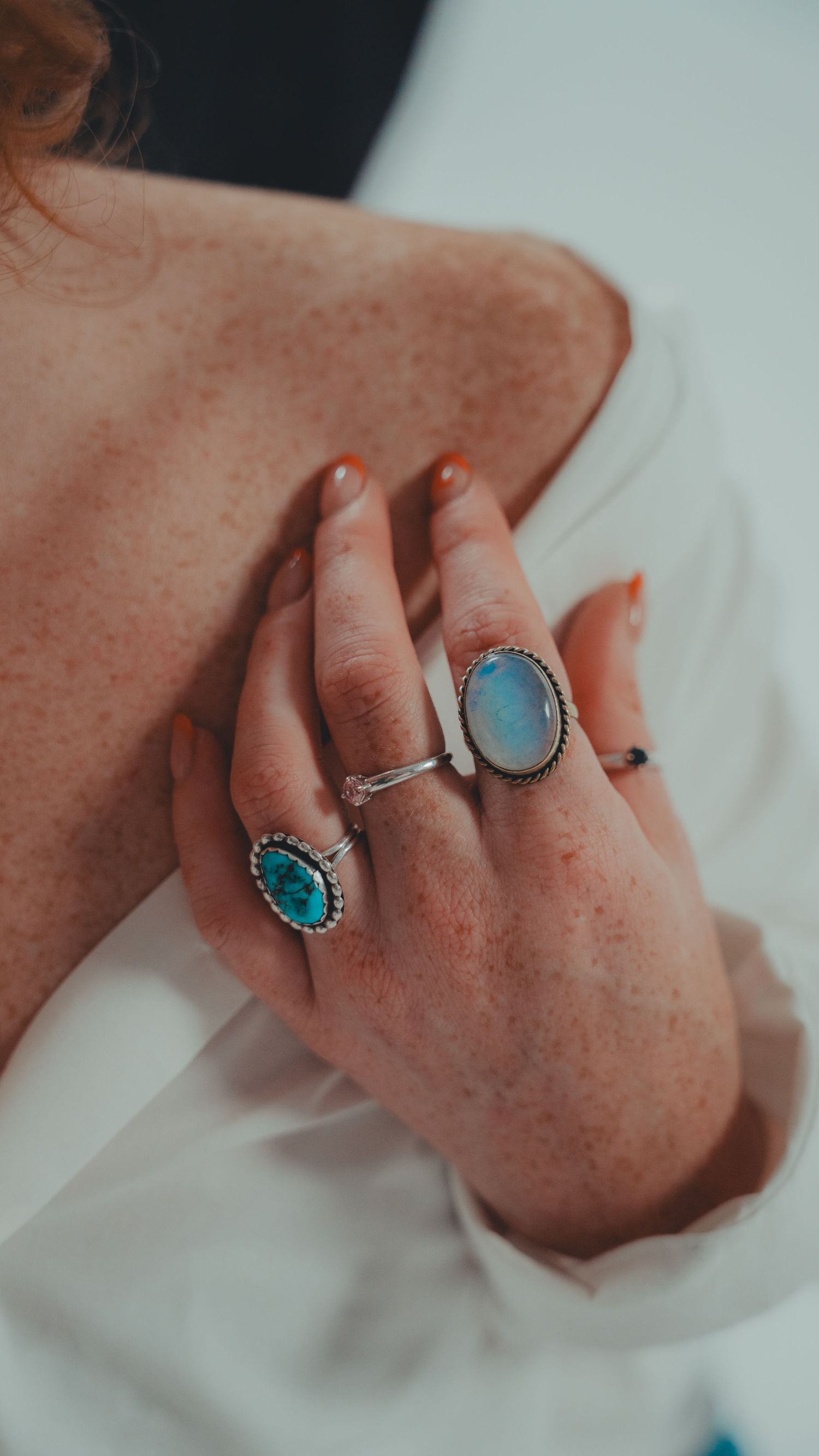 Freckled hand with orange nails wearing turquoise and moonstone silver rings on white fabric.
