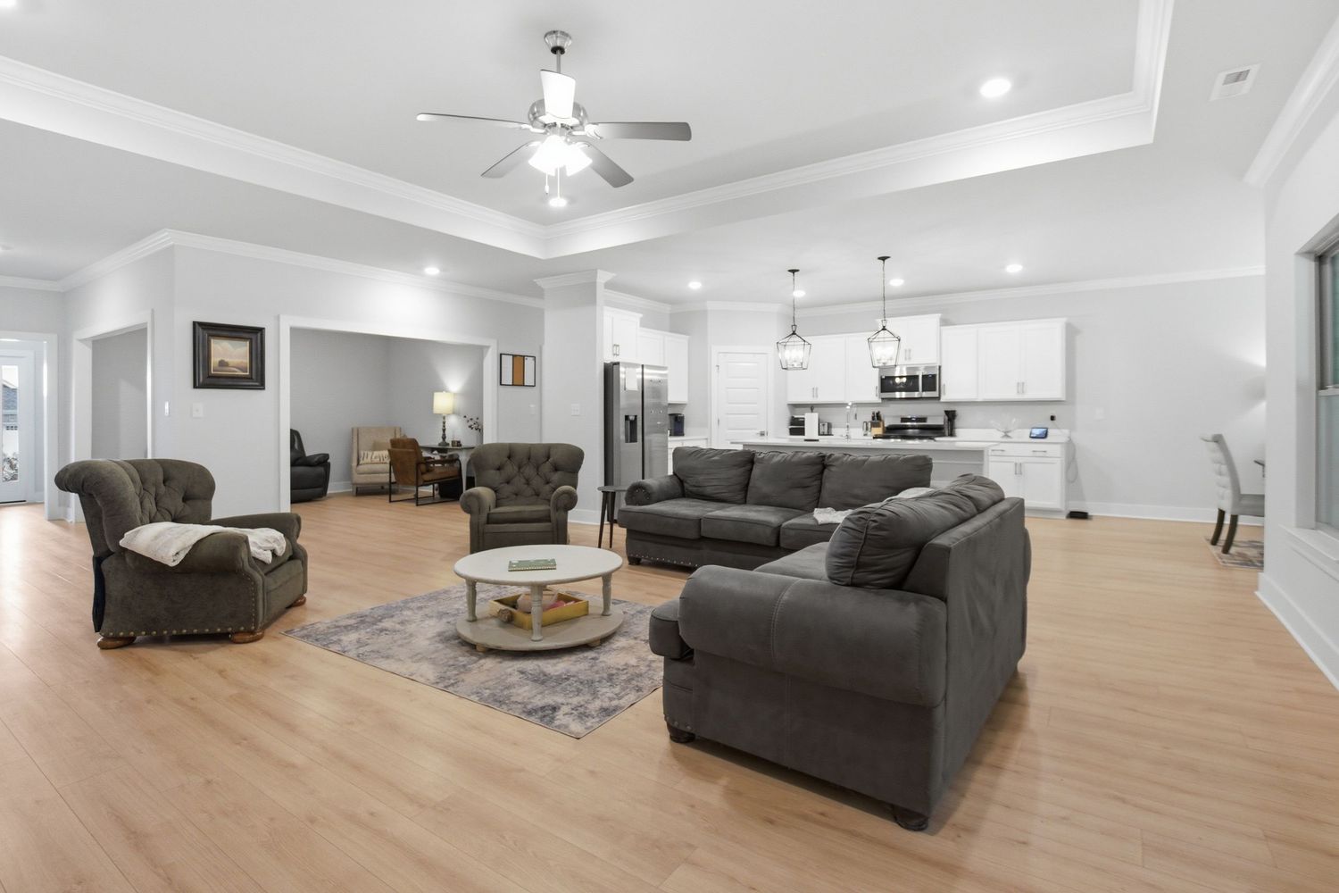 Spacious open-concept living room with hardwood floors, gray sectional sofa, tray ceiling, and modern kitchen visible in background.