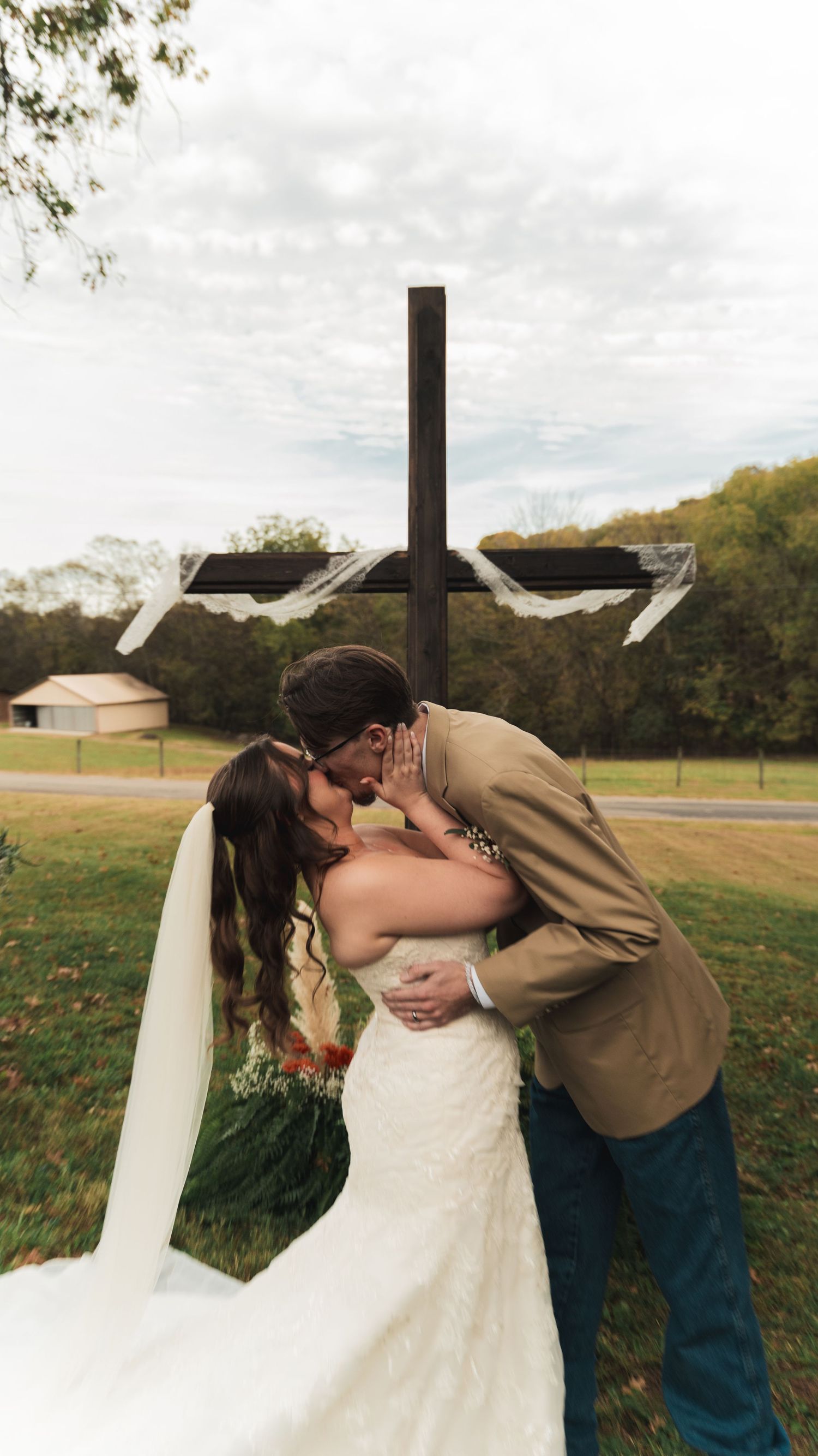 Bride and groom share a romantic kiss in front of a wooden cross outdoors on their wedding day.