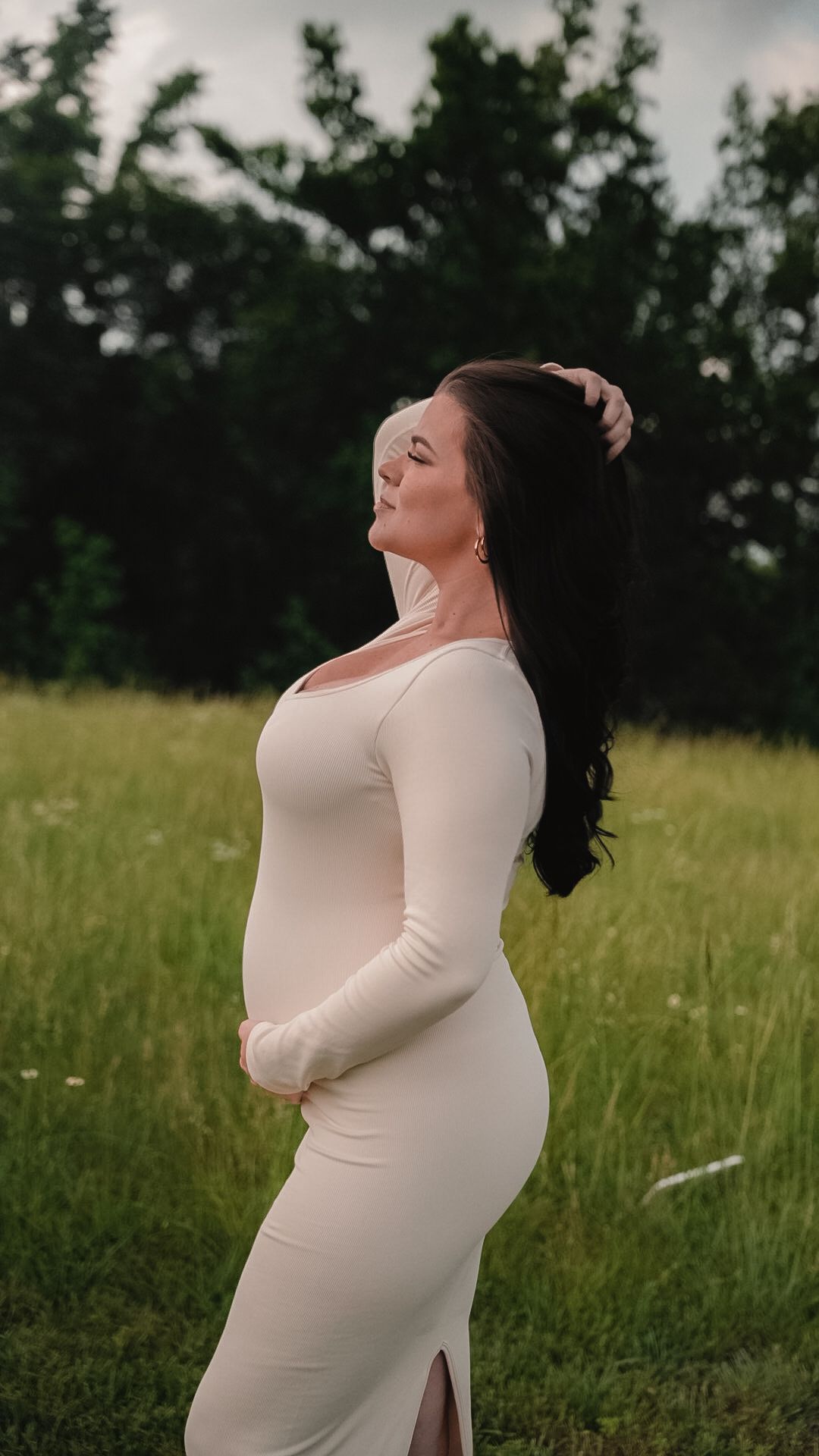 Pregnant woman in white long-sleeve dress poses gracefully in a green field at dusk.