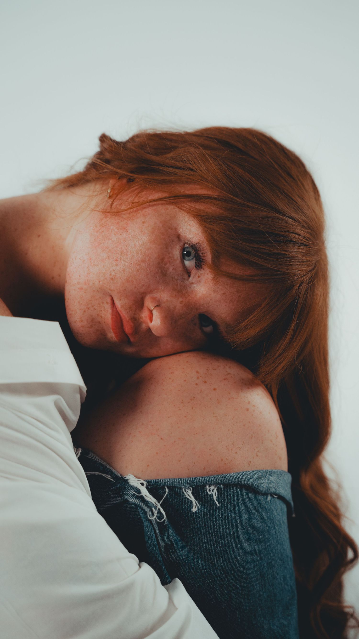 Young woman with red hair and freckles resting her chin on her knee, wearing a white top and distressed jeans.