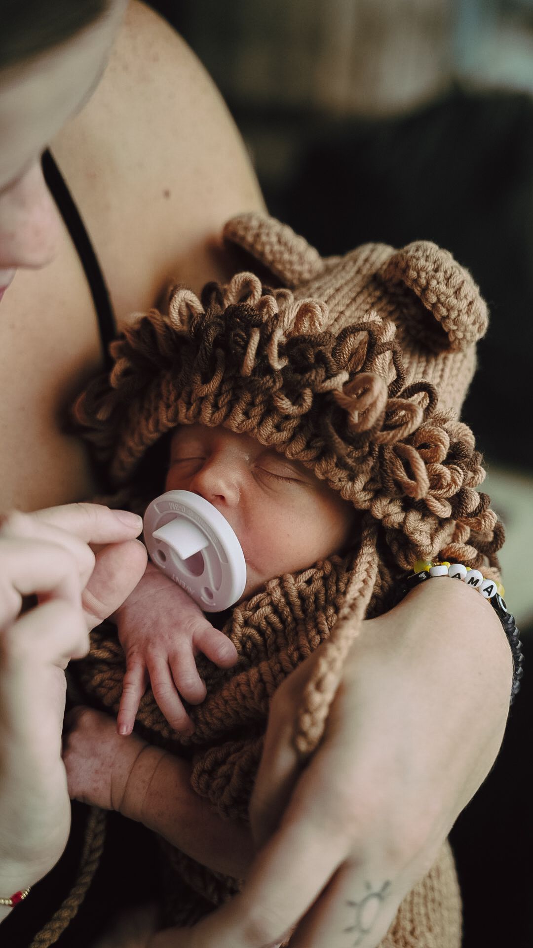 Newborn baby wearing a brown knit bear hat with pacifier, cradled in mother's arms.