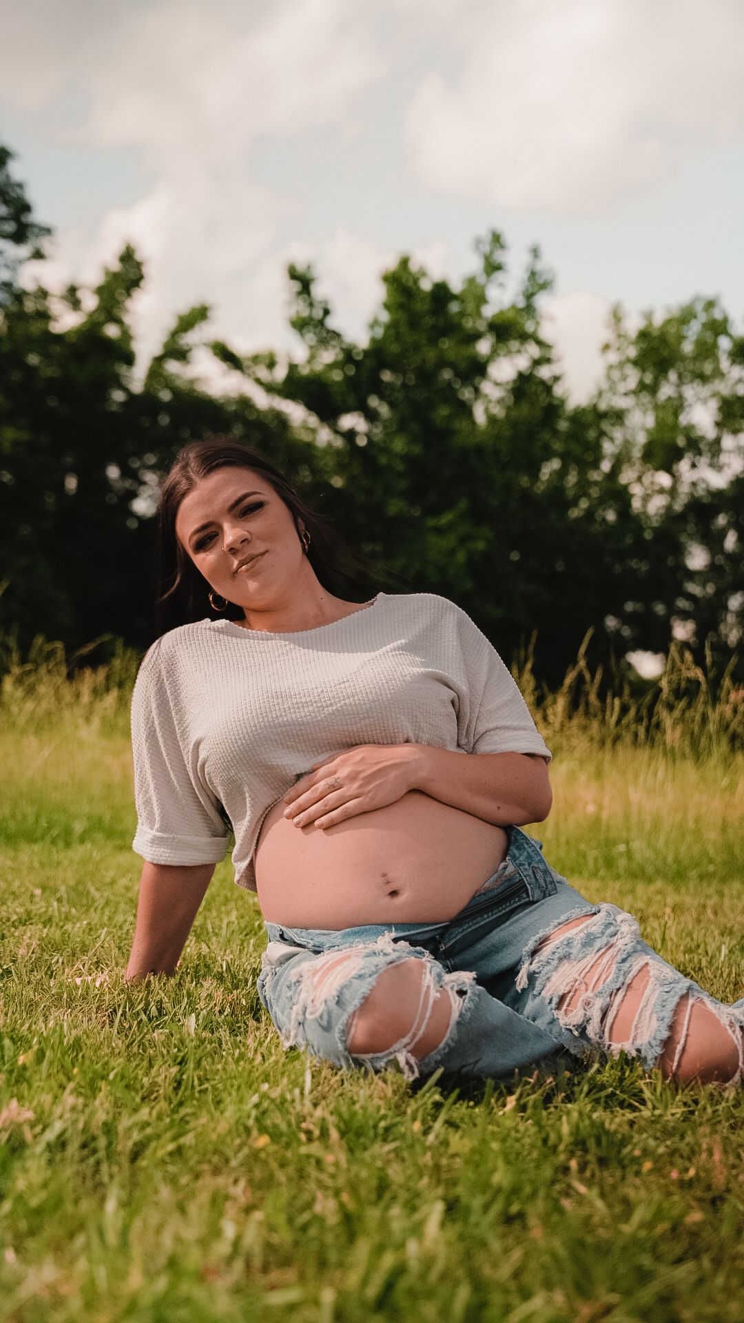 Pregnant woman in white top and ripped jeans sits on grass cradling baby bump with trees behind.