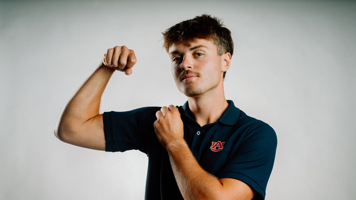 Young man in navy Auburn polo shirt flexing bicep muscle, posing against light gray background.