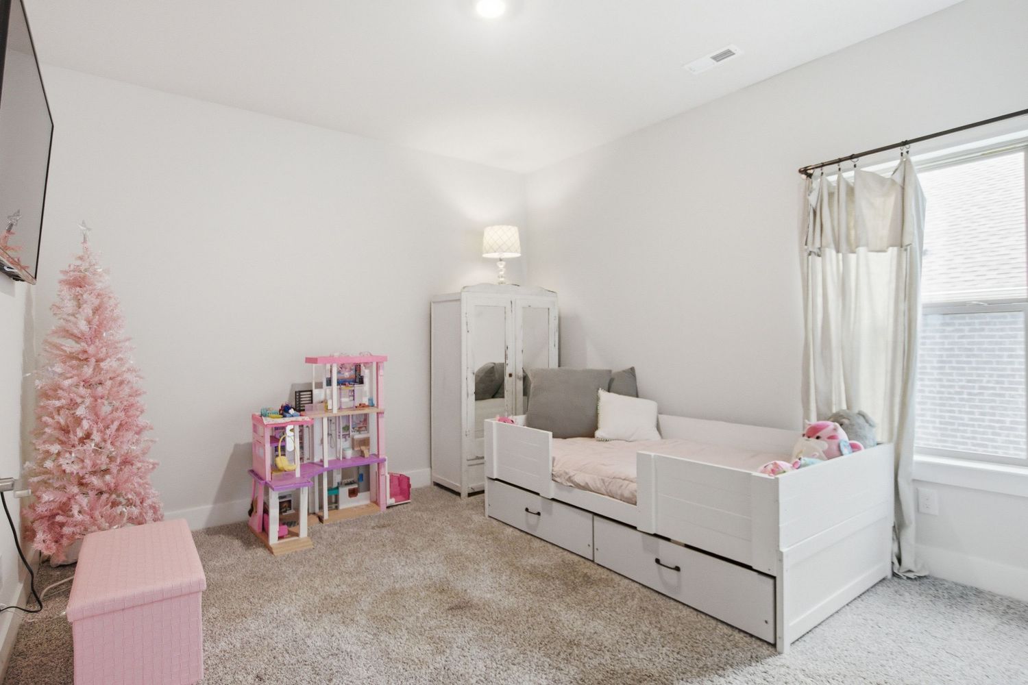 Cozy carpeted kids bedroom with white daybed with trundle, pink Christmas tree, and bookshelf with toys near bright window.