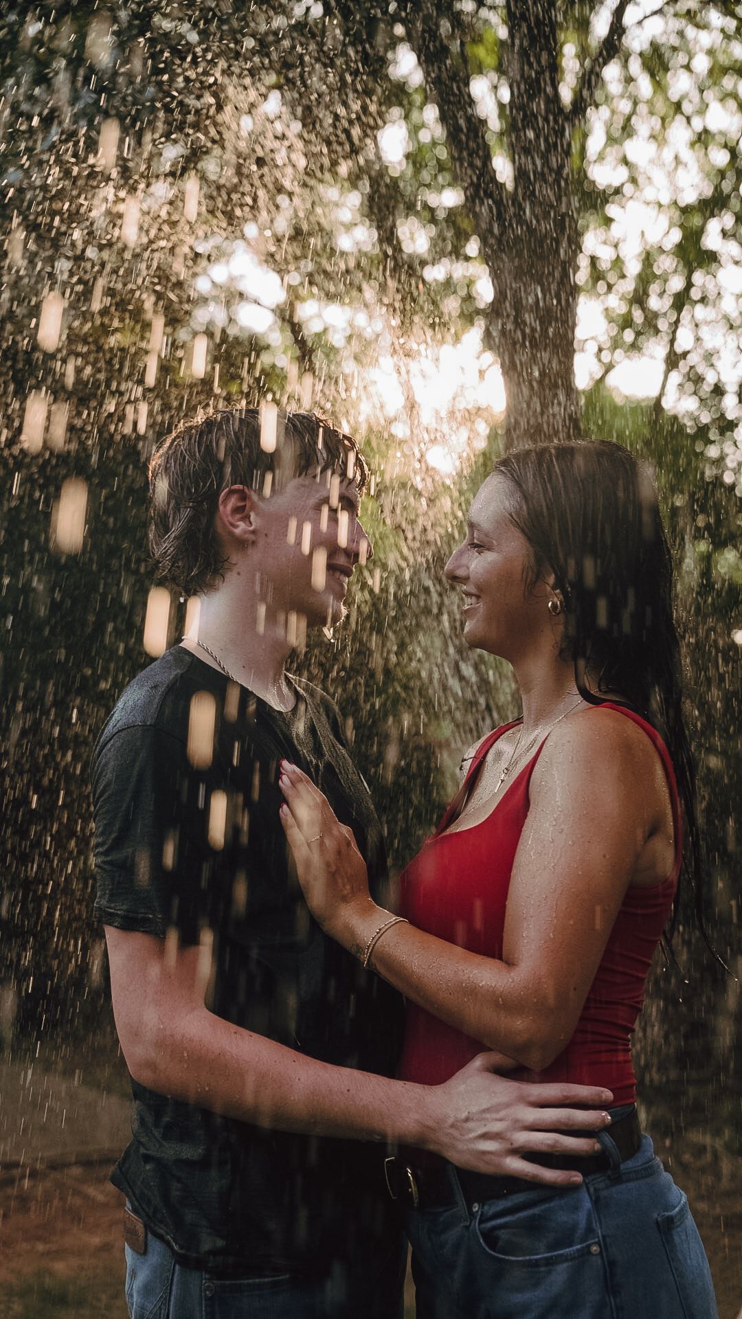 Couple embracing and smiling at each other in the rain outdoors, surrounded by lush green trees.