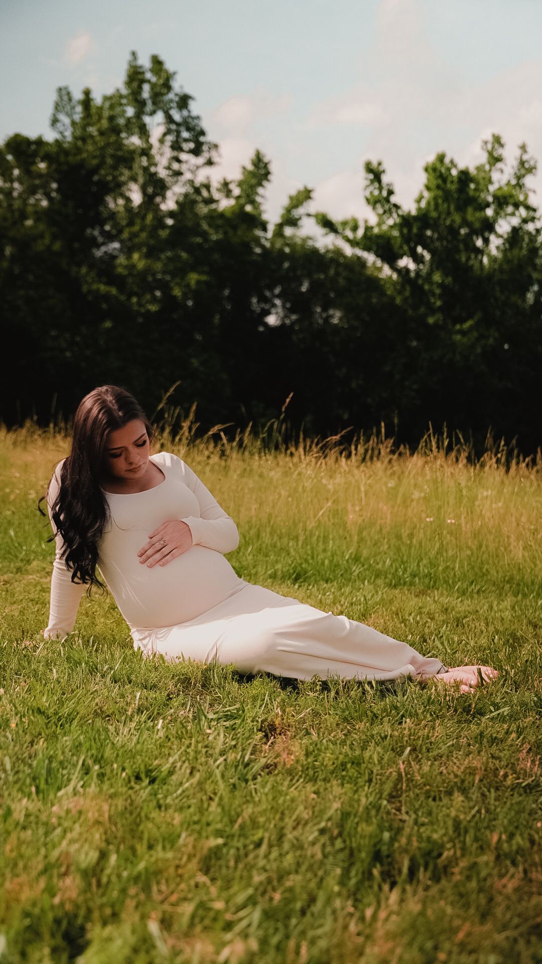 Pregnant woman in white outfit sitting on green grass in a sunny park, gently cradling her baby bump.